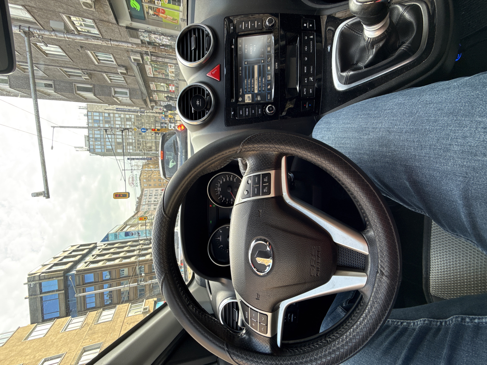 The image shows a view from inside a car, specifically focusing on the driver's perspective. The steering wheel is prominently centered, displaying the logo of the Great Wall Motor (GWM) brand. The dashboard, featuring air vents, a radio display set to FM Radio 1, and various control buttons, are also visible. The driver is wearing blue jeans, partially visible in the lower right area of the image. Outside the car, the scene depicts an urban environment. Buildings of varying heights and architectural styles line the street, including one with a "ДОСТАВКА" (Delivery) sign and a large advertisement. The buildings are a mix of older, Soviet-era designs and more modern structures. There is a traffic light visible, and other vehicles are present, indicating the scene is a street in the city of Sofia, Bulgaria.