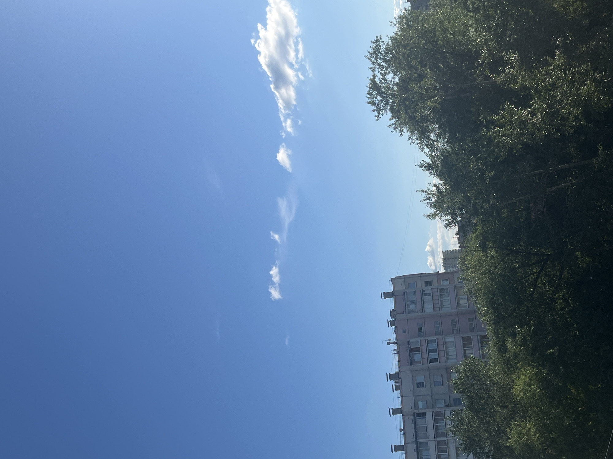A bright, sunny day with a clear blue sky is depicted in this photograph taken in Chelyabinsk, Russia. The image is oriented vertically, with the sky dominating the left half and a building and trees on the right. Scattered white clouds drift across the vast expanse of the sky.
On the right side of the frame, a multi-story apartment building stands, its facade a pale pink color. The building features numerous windows and balconies. The architecture suggests it's a typical residential building found in urban Russia. Towering trees with lush green foliage partially obscure the building, adding a natural element to the urban landscape. The sunlight filtering through the leaves indicates it is daytime, likely late morning or early afternoon, given the brightness. The scene conveys a sense of everyday life in a city, with a focus on the interplay between man-made structures and nature under a clear sky. There is no visible text in the image.