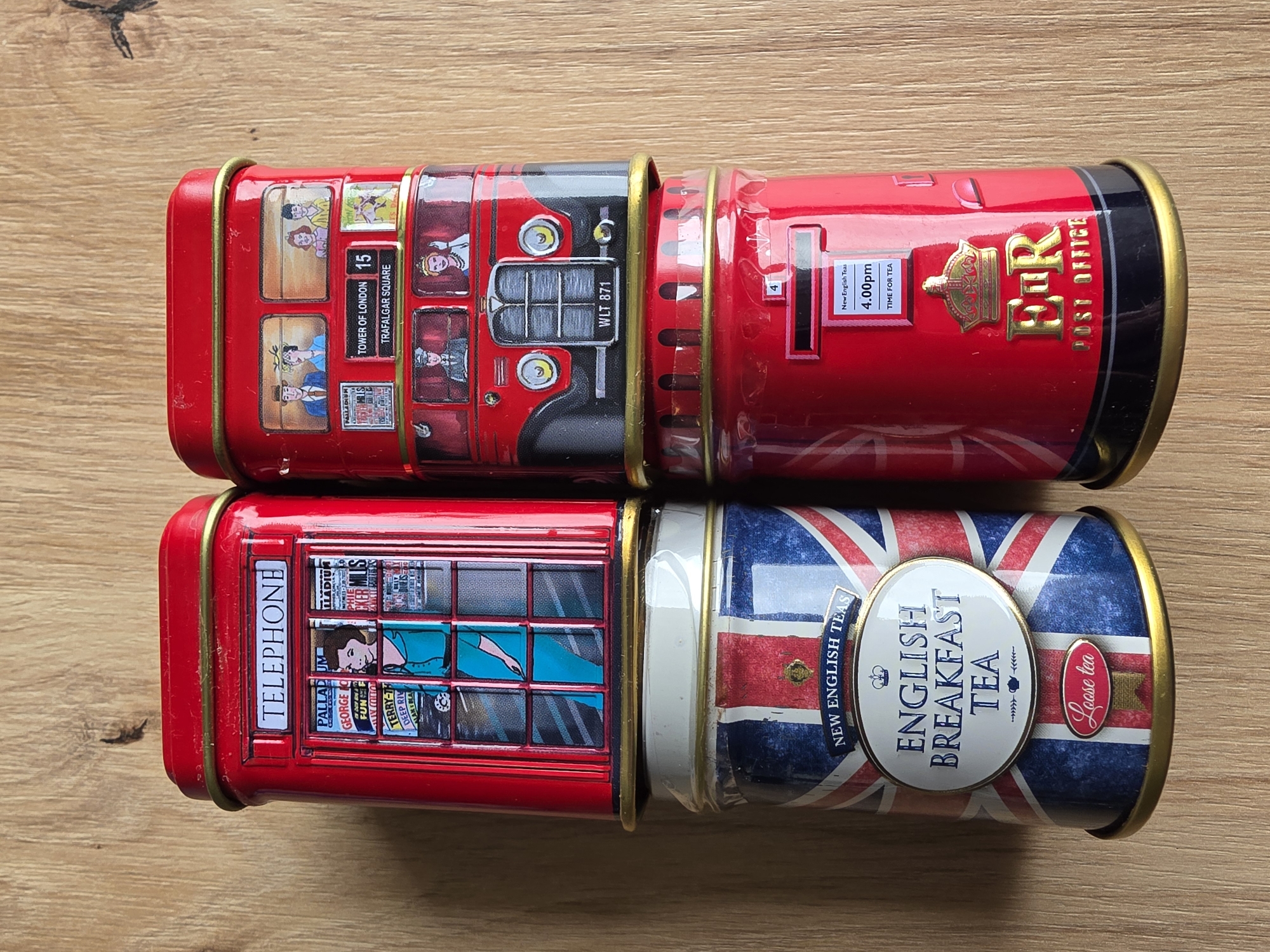 The image shows four cylindrical metal tea tins stacked vertically on a wooden surface. The two tins on the left are stacked one on top of the other, as are the two tins on the right. The overall arrangement forms a rectangular prism.
The top left tin is red and shaped like a British telephone box. The front of the tin features the word "TELEPHONE" in white text at the top. Below this, a window depicts a woman in a teal dress, with various posters visible behind her. The top right tin is also red and designed to resemble a double-decker bus. The front of the tin shows illustrations of people on the upper and lower decks of the bus. A license plate reads "WLT 871".
The bottom left tin is covered in the British Union Jack flag. An oval label in the center of the tin reads "ENGLISH BREAKFAST TEA" in black text, with a crown above and a teapot below. A smaller red oval at the bottom of the tin says "Loose tea".
The bottom right tin is red and shaped like a British post box. It has a small rectangular slot for mail and a label that reads "New English Teas 4.00pm TIME FOR TEA". Below this, a golden crown and the royal cypher "ER" are visible, along with the text "POST OFFICE".
The background is a wooden surface, likely a table or floor, with a visible wood grain pattern. The lighting suggests an indoor setting. There are no people or animals present in the image.
There is no information within the image or its visual context that suggests a connection to the city of Poprad, Slovakia.