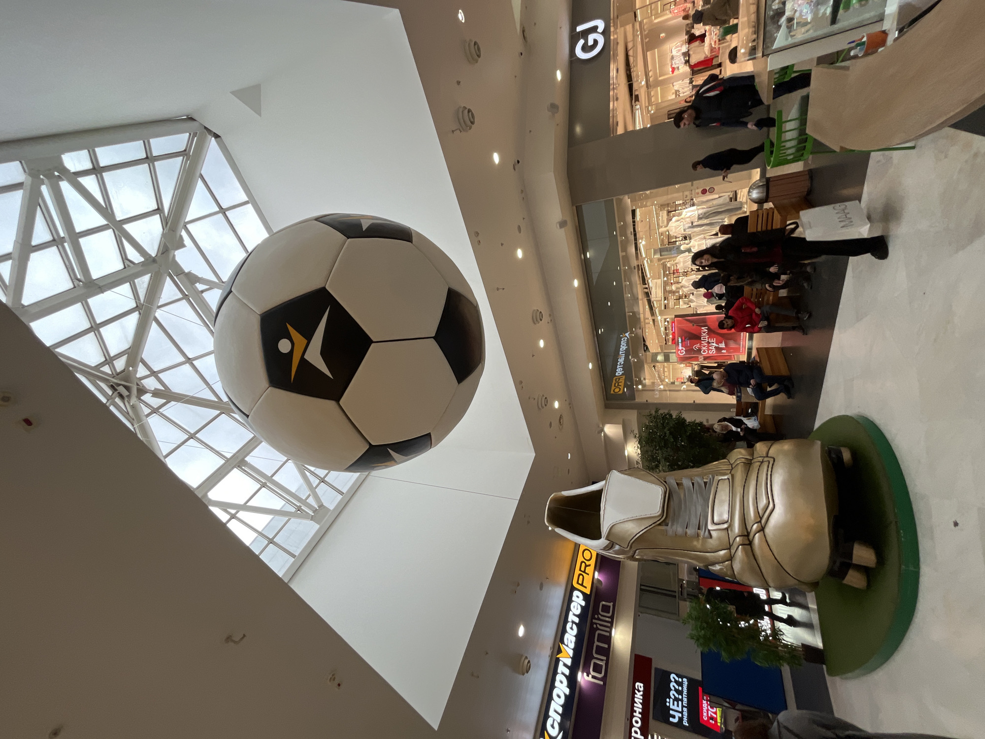 The image is taken from inside a shopping center in Krasnoyarsk, Russia. A large soccer ball is suspended from the ceiling near a windowed skylight. Below the soccer ball, a large golden soccer shoe statue is visible in front of a series of retail storefronts, including “Sportmaster PRO Familia.” People are walking, sitting, and shopping on the floor above. Visible signage includes “GJ” and “MAAG.” Text also written on the storefronts includes phrases such as “Black Friday,” "скидки," and "SAVE." The image is taken from a ground-level, upward-angled perspective.