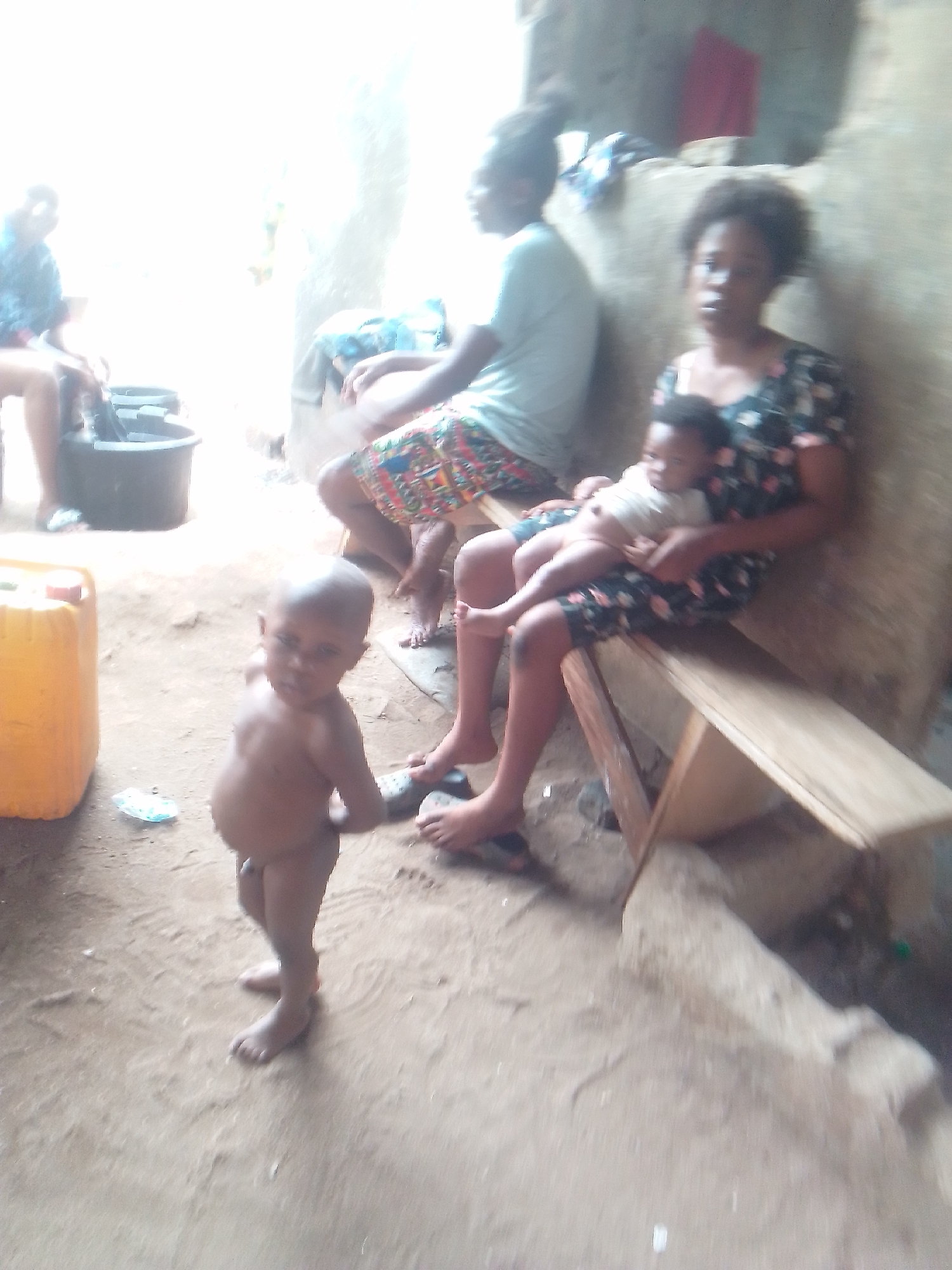 The image shows a group of people in what appears to be an outdoor or semi-outdoor setting in Owerri, Nigeria. A young, bare-chested child stands in the foreground, looking towards the camera. To the right, a woman sits on a simple wooden bench holding a baby in her lap. Another woman sits behind them, and a fourth person is partially visible in the background to the left. A yellow jerrycan is visible to the left. The ground is dirt or sand. The setting looks like a modest or rural environment.