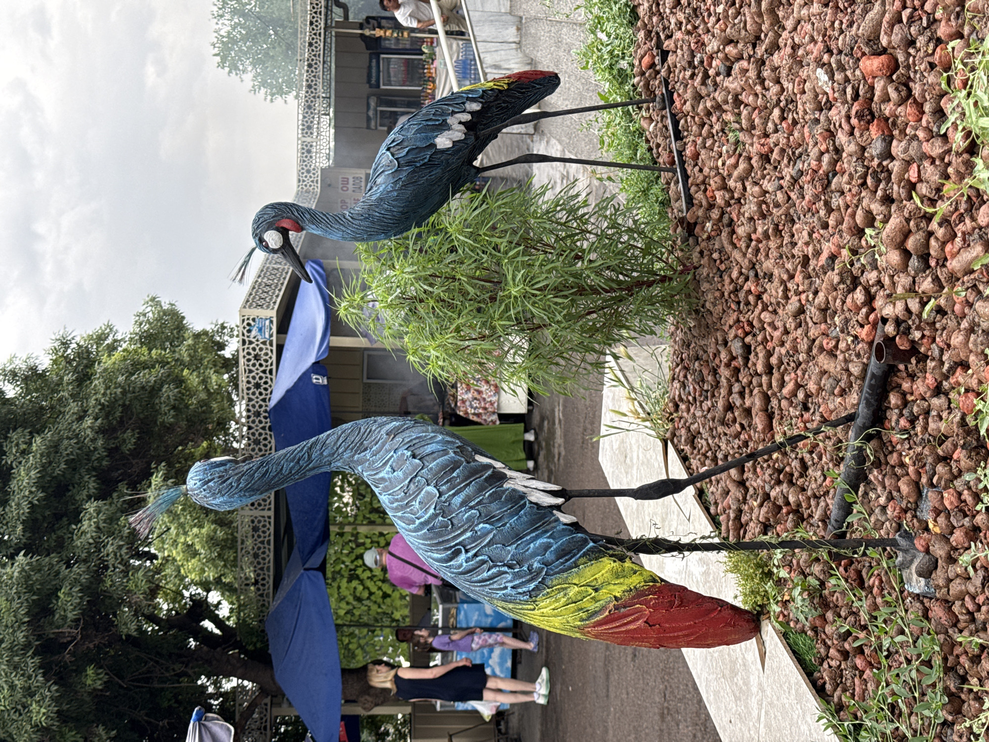 The image shows two ornamental crane sculptures standing in a pebbled area in a public space, possibly a market or park, in Tashkent, Uzbekistan. The cranes are painted with blue, white, yellow, and red colors. A small green shrub is positioned between the two cranes. The background includes trees, a blue tent, people walking around, and vendor stalls, which are typical of a busy outdoor market scene. The sky is overcast.