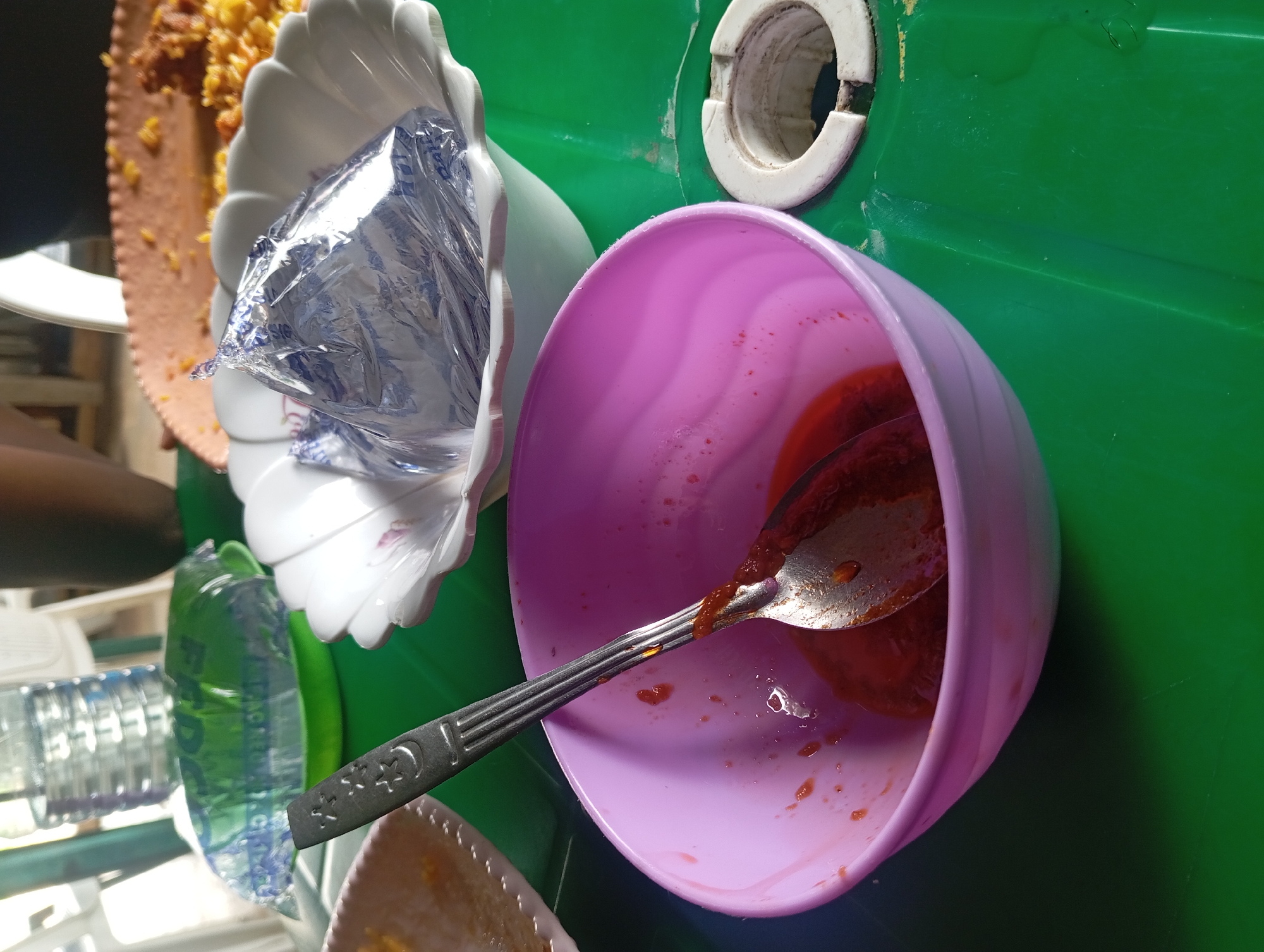 The image depicts a close-up, slightly elevated view of food items on a green table. In the foreground, a pink bowl contains a reddish sauce and a spoon. To the right of the bowl, a white cylindrical object with a hole in the center is visible. Behind the pink bowl, a white, fluted bowl holds an object wrapped in shiny, silver foil. Further in the background, a green plastic tray is partially visible, and a bottle of water stands in the upper left corner. A plate with yellow rice and some other food is in the upper right background. The scene appears to be set indoors, likely in a casual dining environment.