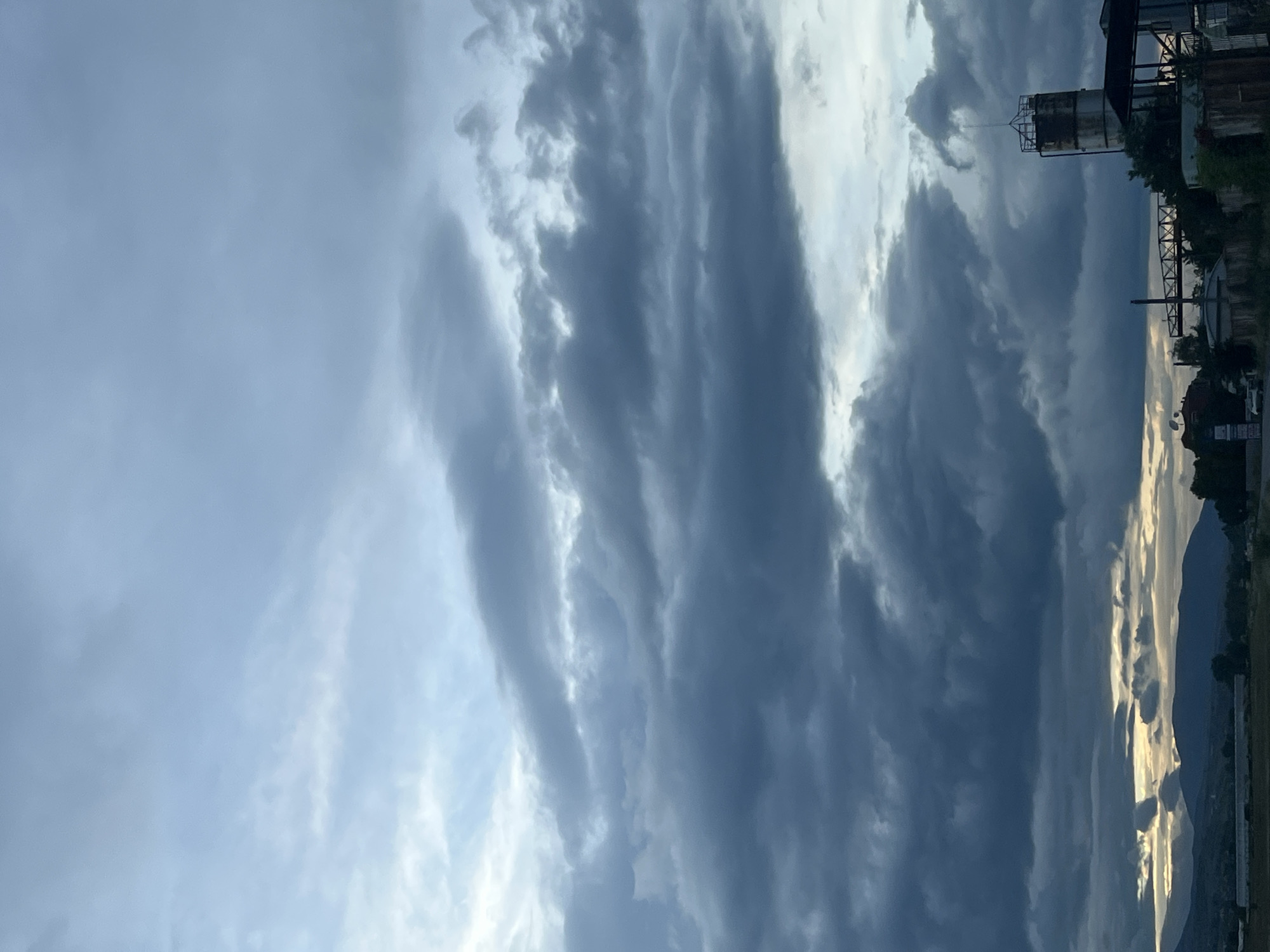 The image depicts a sky filled with dark, striated clouds. A structure resembling an industrial tank is visible against the clouds. In the distance, the outline of a mountain range is discernible. The photo was taken near a city, and according to the metadata provided, it is located in Blagoevgrad, Bulgaria.