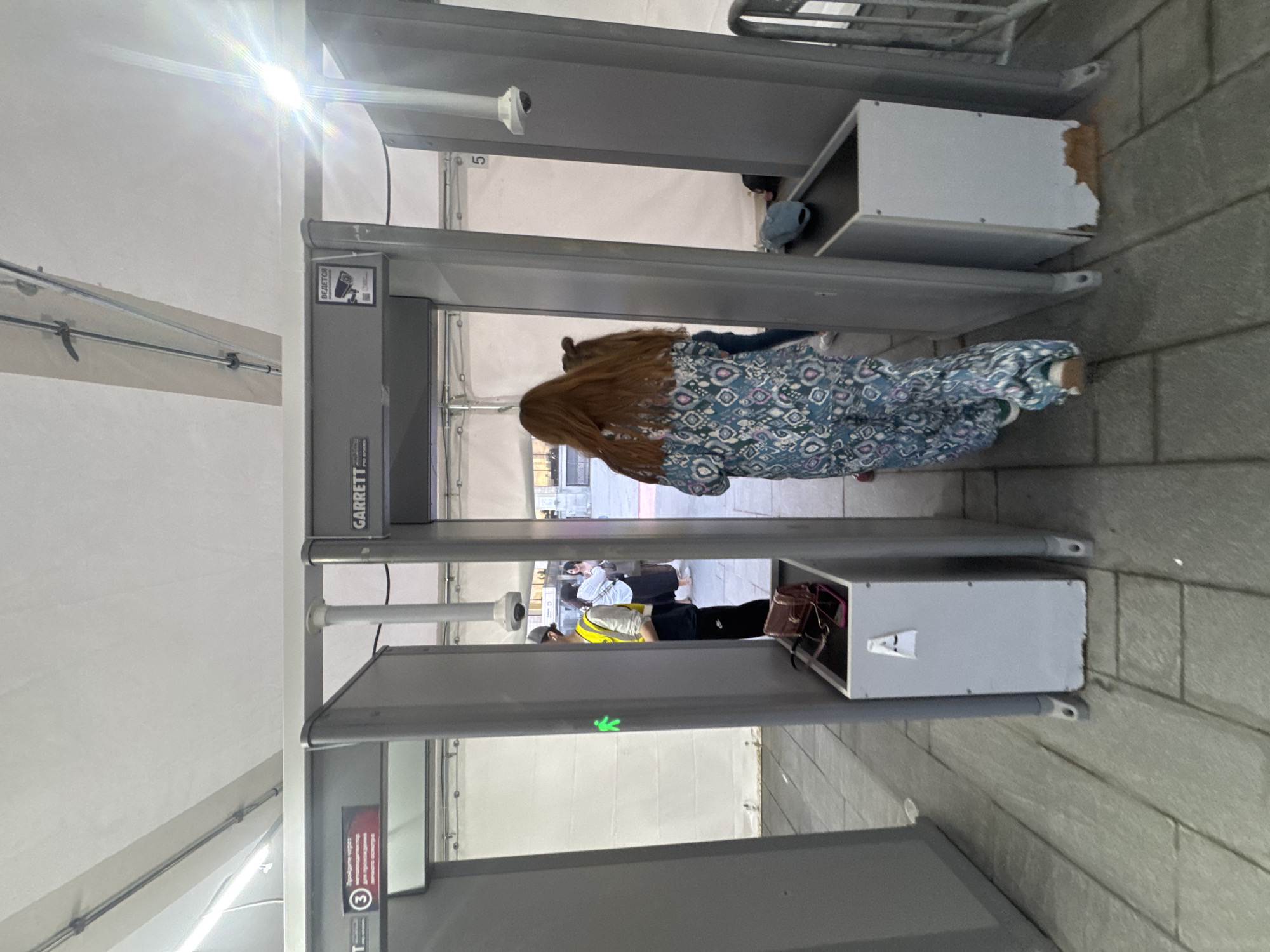In Moscow, Russia, at a security checkpoint, a woman with long auburn hair, wearing a blue and pink patterned jumpsuit, walks through a metal detector. She is followed by another person, whose face is not visible. A security guard in a yellow vest and cap stands to the left of the detector, facing away from the camera. In the foreground, a metal bin contains a purse and two cell phones. The environment appears to be outdoors, with a paved ground surface and a white tent-like structure in the background. The metal detector is a Garrett brand, and a surveillance camera is visible above the detector on the right.