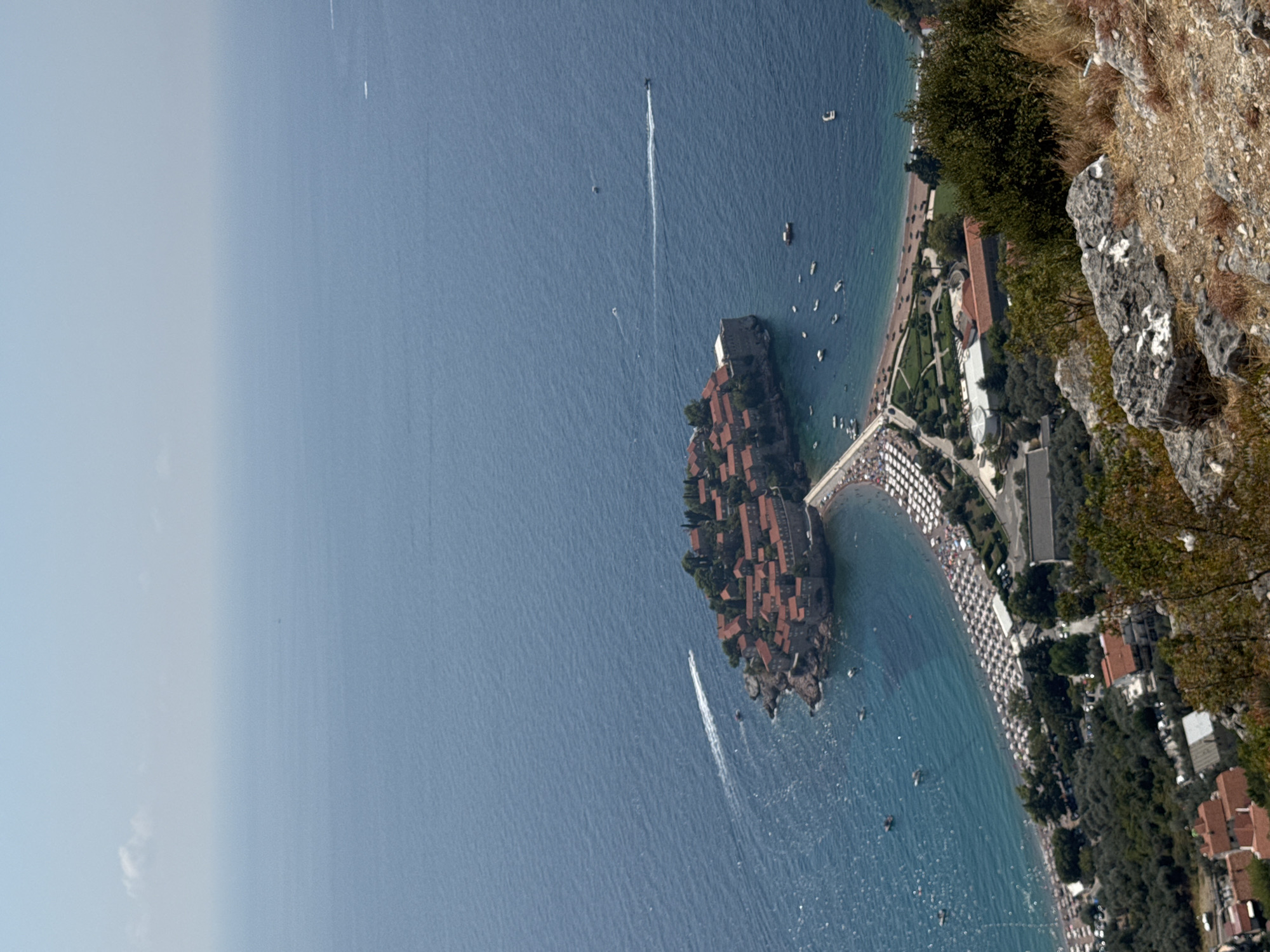 This is an aerial photograph of Sveti Stefan, Montenegro, taken from a high vantage point. The image captures a peninsula town jutting out into a vibrant blue sea. The town itself is a cluster of red-tiled buildings, tightly packed together on a small rocky island, connected to the mainland by a narrow causeway. The causeway is lined with rows of white beach umbrellas, indicating a populated beach area. Beyond the beach, a road runs parallel to the coastline, with cars and buildings visible. The sea is dotted with several small boats and a few boats are creating white wakes as they speed across the water. In the foreground, rocky terrain with sparse vegetation and dry grass suggests the elevated viewpoint. The sky is a hazy, pale blue, with a few wispy clouds. The overall scene depicts a picturesque coastal resort town on a clear day.