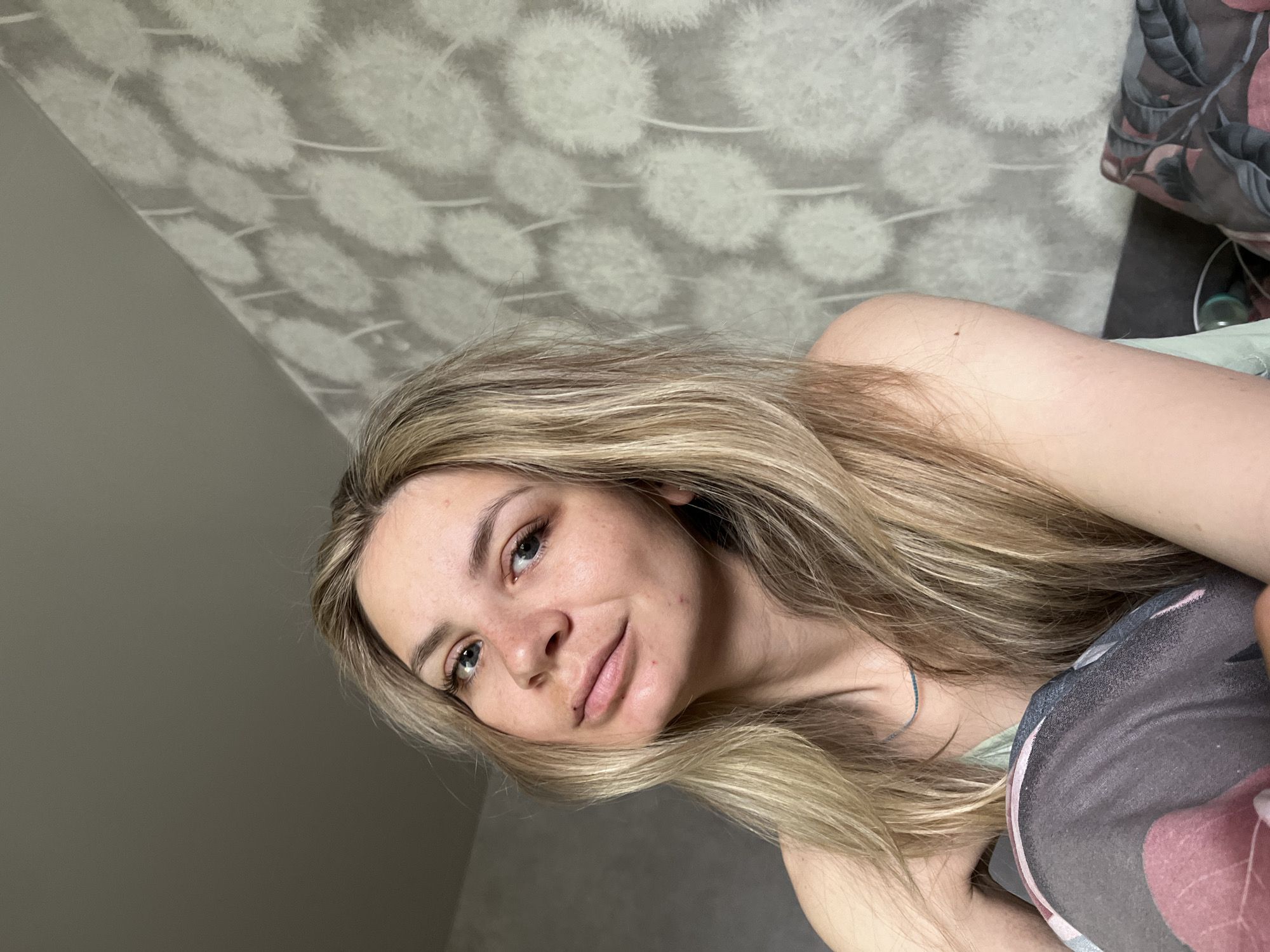A woman with long, blonde hair is lying in a bed. The background consists of a grey and white patterned wall and a bedspread with a floral design. The location appears to be indoors. There is no specific information in the image to verify the location in the city of Chaikovsky, Russia.