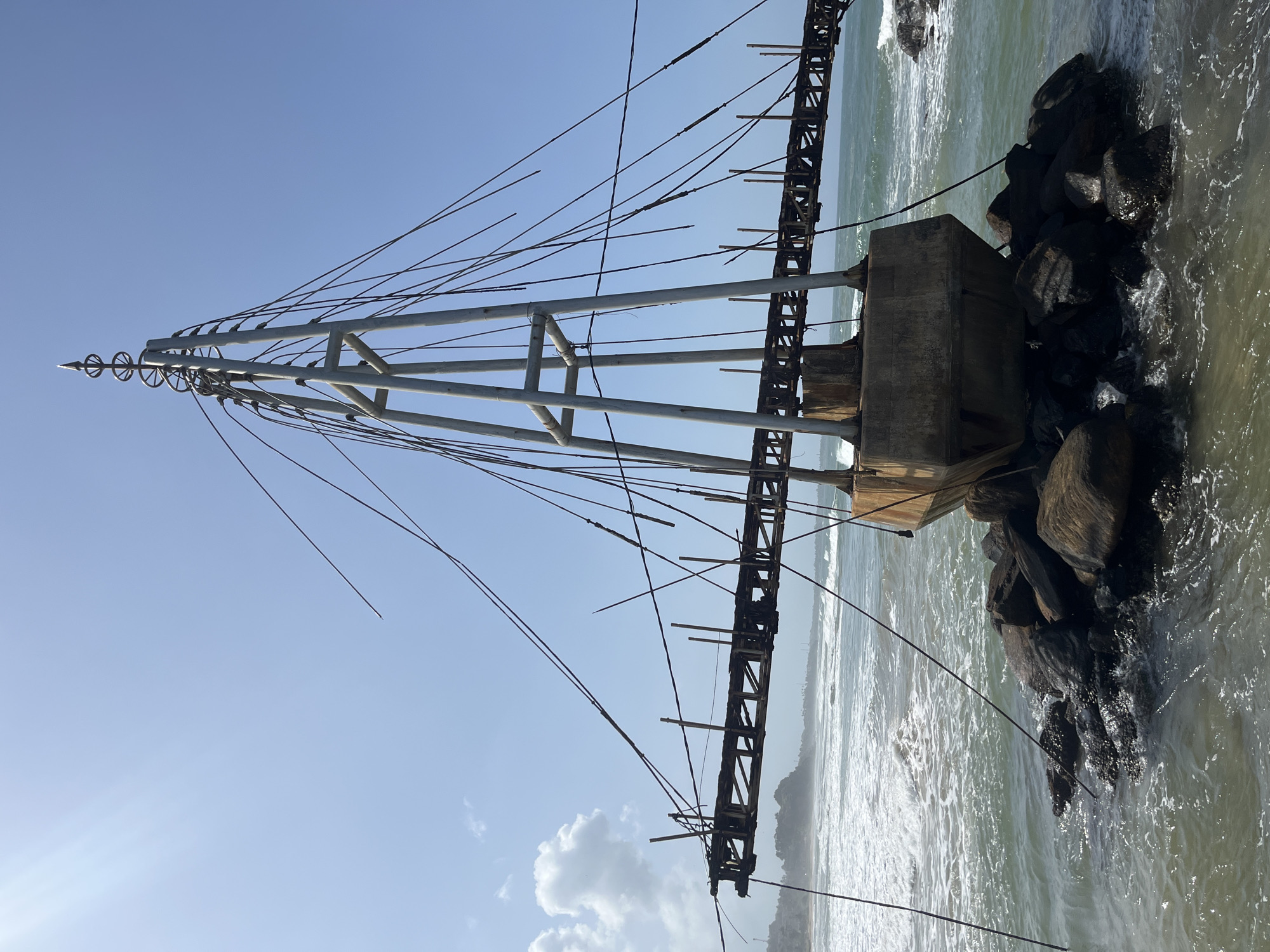 The image depicts a skeletal metal structure with a concrete base resting on large rocks in the ocean, near the shore. The structure appears to be a lighthouse or a navigational marker, possibly in a state of disrepair. The sky is clear and blue with a few wispy clouds. The ocean has moderate waves crashing against the rocks and the shore. The scene is likely located in Matara, Sri Lanka, a coastal city known for its scenic beaches and historical landmarks. The image does not contain any people.