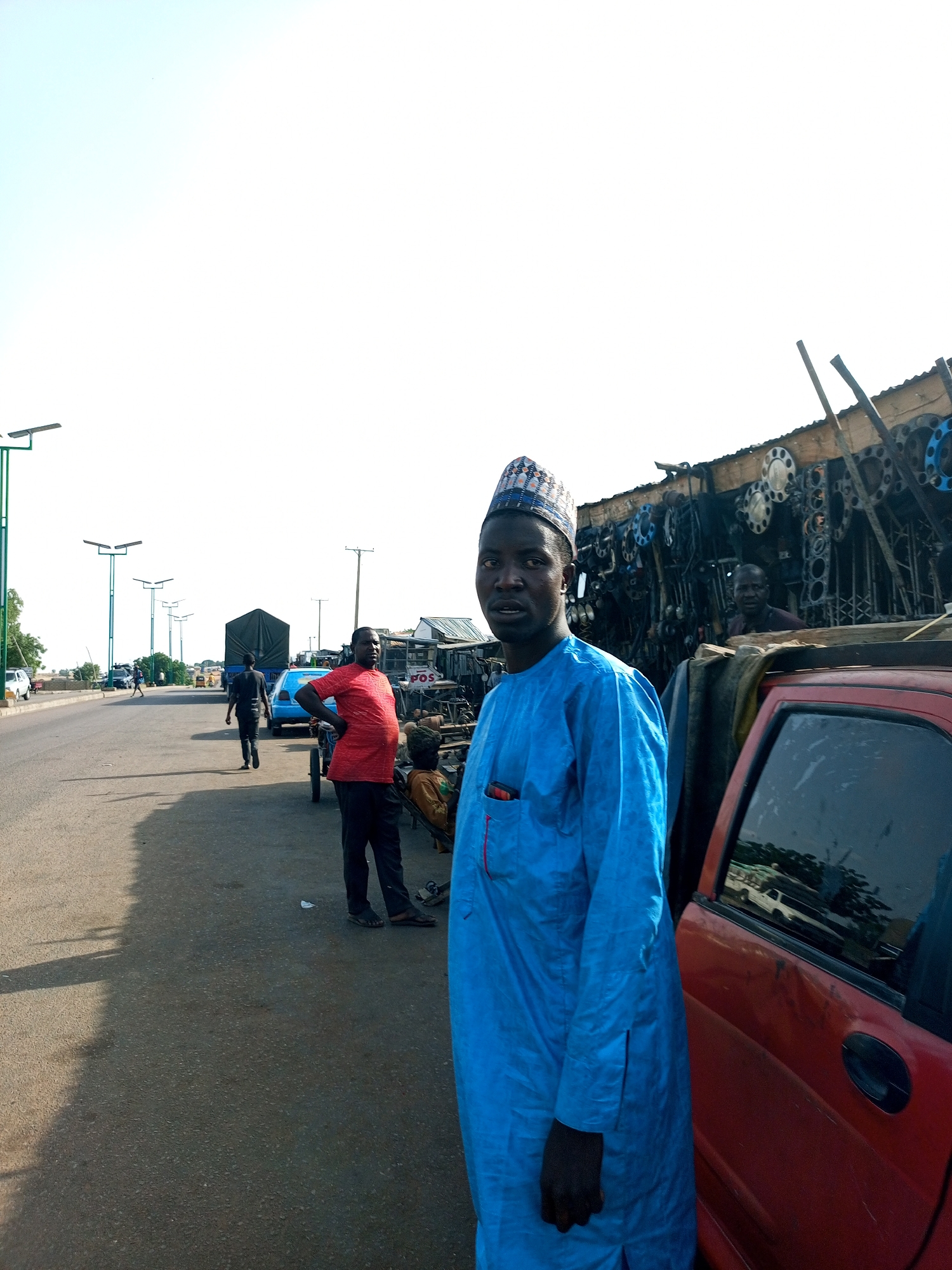 The image depicts a roadside scene in Gombe, Nigeria, likely an informal marketplace or repair area. In the foreground, a man wearing a blue kaftan and a patterned cap stands near a red vehicle. Behind him, a stall displays a variety of mechanical parts, possibly for vehicles, with several metal objects resembling gears or flanges visible. To the left, a paved road stretches into the distance, lined with streetlights. Several people are visible on the road, including a man in a red shirt standing with his hands on his hips, and others walking or standing near vehicles and carts. The environment suggests a busy, open-air commercial area. The sky is bright and clear.