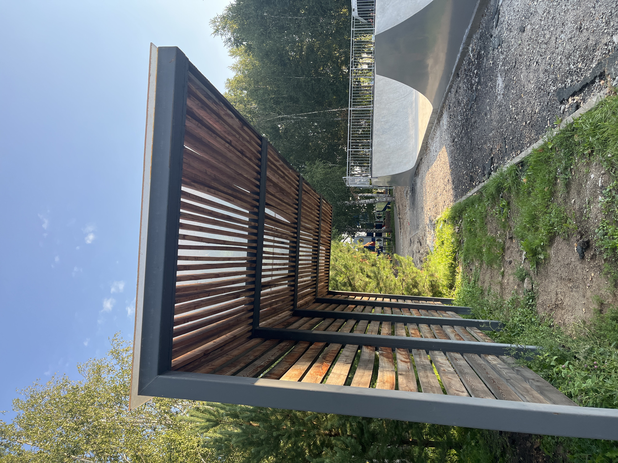 In Novye Turbasly, Russia, an outdoor public space features a modern wooden slatted canopy extending over a pathway adjacent to a concrete skate park. The canopy has a dark grey metal frame and a translucent roof allowing sunlight to filter through. The wooden slats are oriented vertically and are weathered in appearance. A wooden fence, also with vertical slats, runs alongside the pathway under the canopy, creating a sense of enclosure. Green grass and some small plants grow along the path and at the base of the fence. In the background, a concrete skate park with curved surfaces is visible, bordered by a metal railing. Trees with green foliage form a dense backdrop, suggesting a park or wooded area. The sky is clear blue with a few scattered white clouds. The scene appears to be newly constructed or undergoing some development, with loose gravel and some debris on the ground near the skate park. No people are clearly visible or engaged in activity.