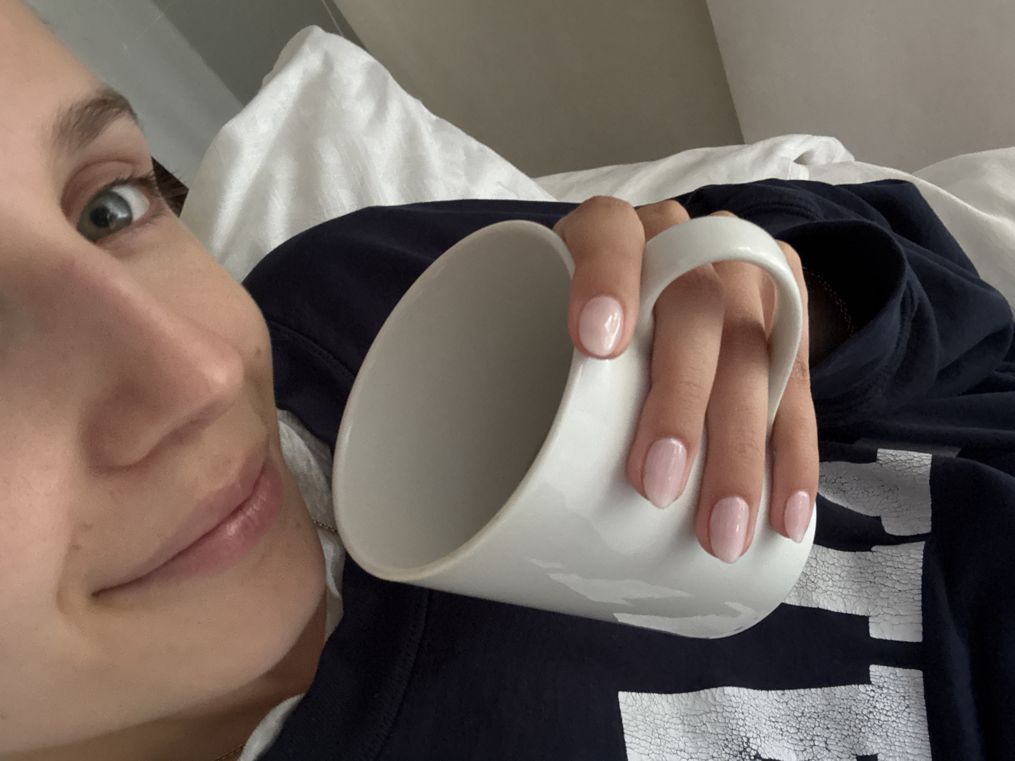 A close-up, slightly angled overhead shot shows a person holding a white ceramic mug. The person's face is partially visible, with their right eye and nose in focus, and their lips are slightly parted in a soft smile. The person has light skin and blue-green eyes. Their fingernails are painted a light pink color. They are wearing a dark blue sweatshirt with a white graphic on the left side. The background consists of white bedding. The image does not contain any information that would allow for identification of a specific location within Ciudad de México, Mexico.