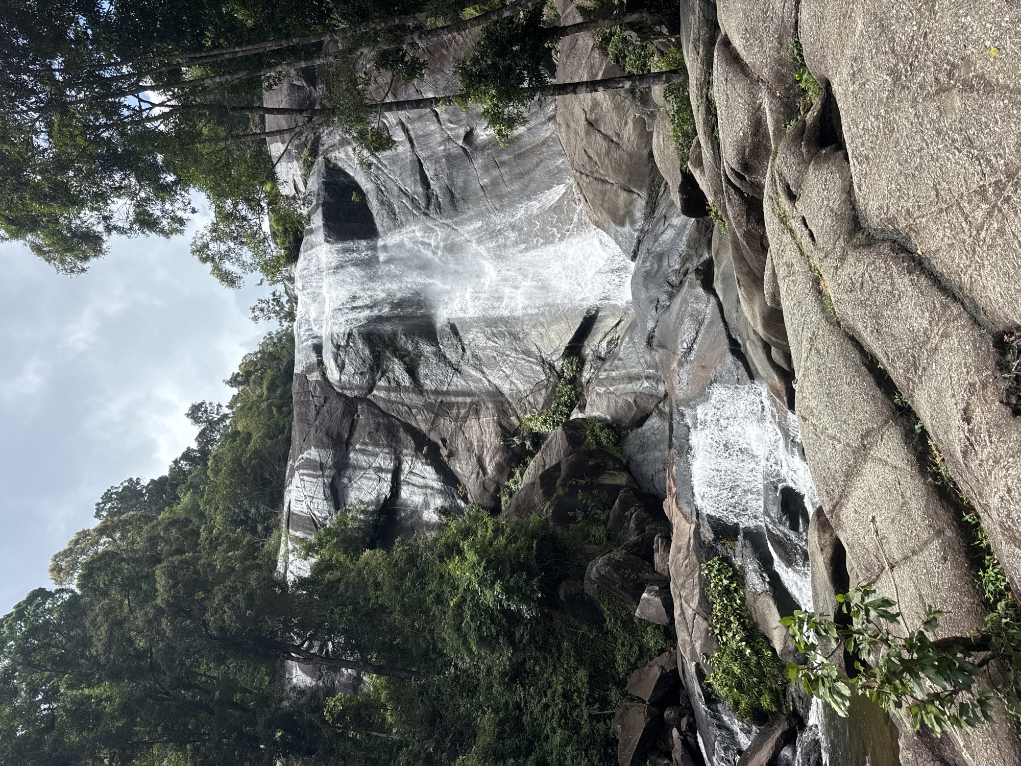 The image depicts a multi-tiered waterfall cascading down a large granite rock face, surrounded by lush green vegetation. The waterfall is wide, with water fanning out and flowing over various rock formations. The foreground is dominated by large, weathered granite boulders. In the background, dense trees and foliage cover the slopes leading up to the top of the waterfall. The sky is overcast with some lighter patches. The scene appears to be a natural landscape, likely a tourist attraction given its picturesque nature. This is a photograph of the Temurun Waterfall located in Langkawi, Malaysia. No people are visible in the image.