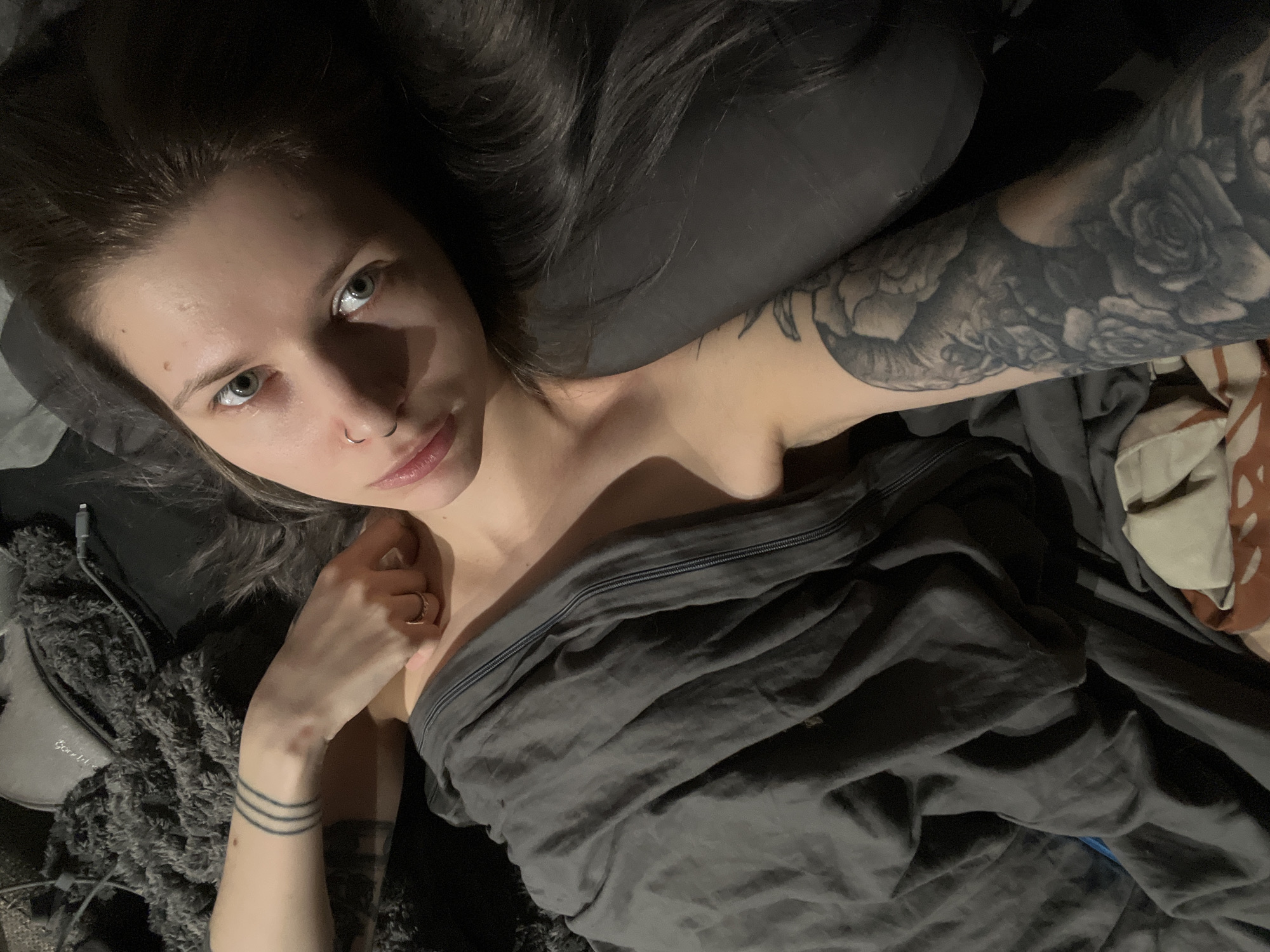 A woman with fair skin, light eyes, and dark hair lies in bed under a gray blanket. She has a nose piercing and tattoos on her arm and wrist. The environment appears dark and intimate. No information related to Khimki, Russia is evident in the image.