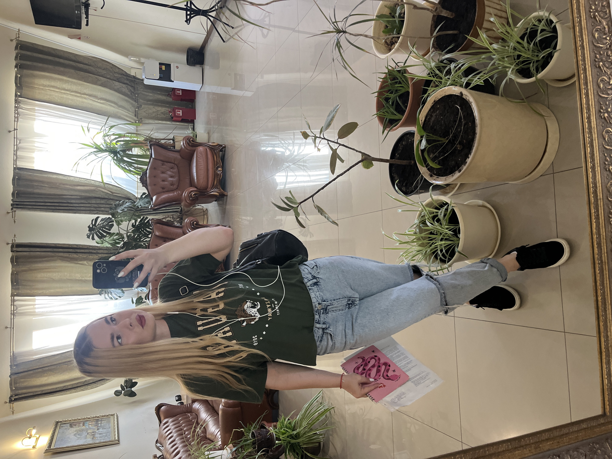 The media file shows a young woman taking a mirror selfie in what appears to be a well-lit room with tiled flooring and large windows. The room contains several potted plants, some of which are large and leafy, suggesting an indoor environment. A leather armchair is visible in the background, as is a framed picture on the wall. The woman is wearing a dark green t-shirt, light blue jeans, and black sneakers. She has headphones in her ears and is holding a smartphone and a pink notebook with a snake design. Her hair is long and blonde, and she is wearing makeup. The overall impression is that of a casual, everyday moment captured in a relaxed setting. The image is vertically oriented, and the lighting suggests it could be daytime. There is text on the woman's t-shirt, but it is difficult to read clearly. The pink notebook has text that is also indistinct. The location context is Krasnodar, Russia.