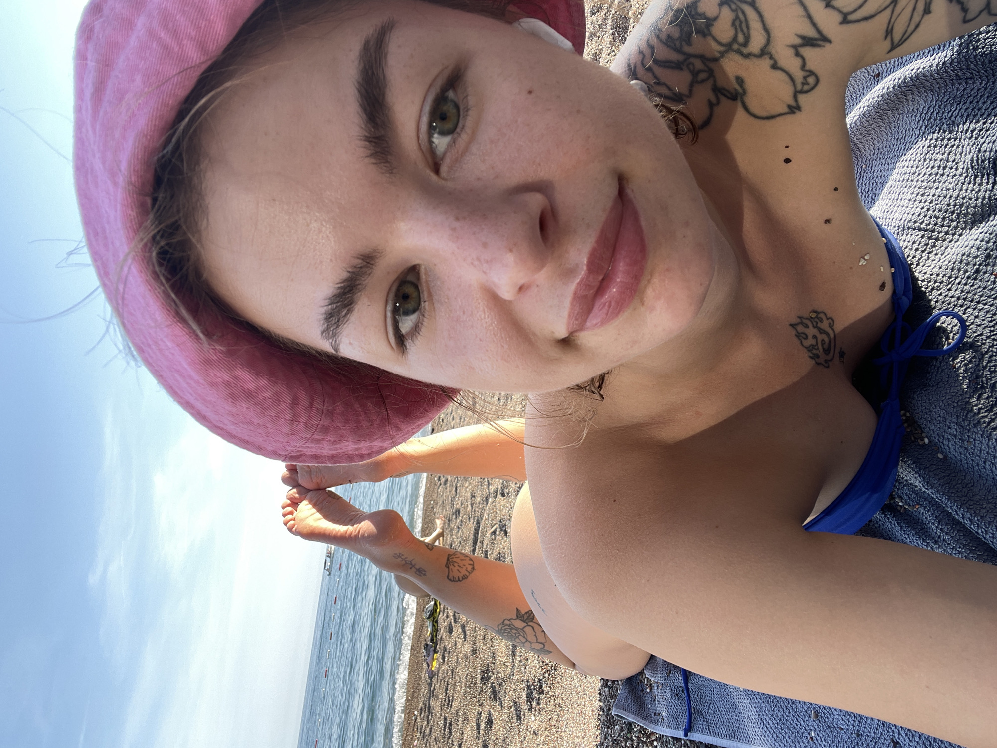A young woman is captured in a close-up, low-angle selfie-style shot, lying prone on a blue beach towel on a sunny beach in Rafailovići, Montenegro. She looks directly at the camera with a subtle smile.
She wears a pink bucket hat, partially covering her light brown hair, and a blue bikini top. White wireless earbuds are visible in her ears. Her face is adorned with numerous freckles, and she has striking green eyes and glossy, full lips.
Her body features several tattoos; intricate designs are visible on her arms and shoulder, while her legs display a seashell motif, a floral pattern, and what appear to be East Asian characters. Small pebbles and grains of sand are scattered on her skin and the blue towel.
In the background, the calm, turquoise sea stretches out under a clear, bright blue sky, with a visible horizon line. Distant buoys or markers are present in the water. The beach itself is pebbly. Her bare feet are bent upwards, also showing tattoos on her ankles and lower legs. The scene conveys a relaxed and joyful atmosphere on a bright day by the sea. No legible text is visible in the image.