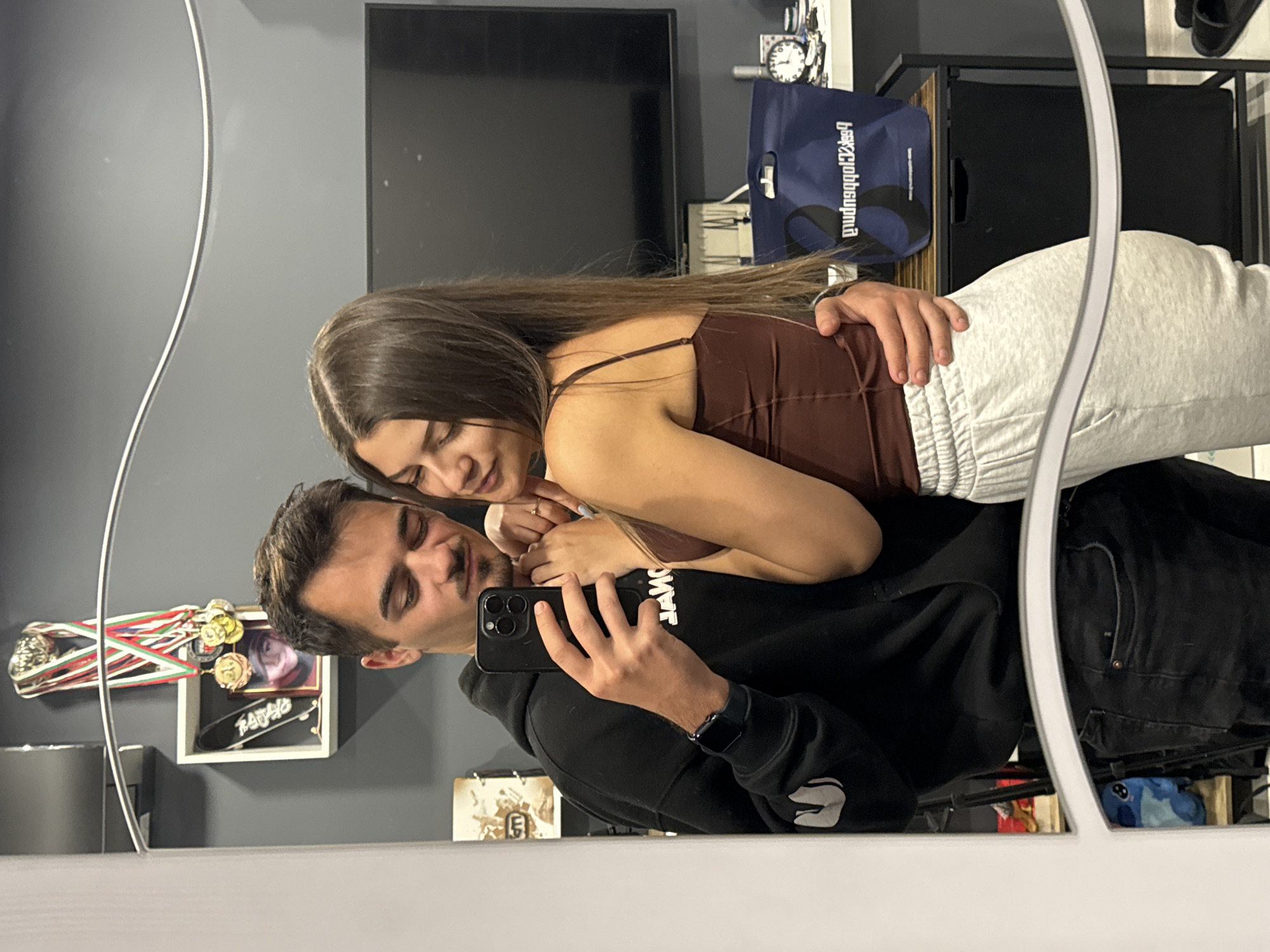 A photograph shows a couple in a mirror selfie in a room in Sofia, Bulgaria. The man, wearing a black hoodie, is holding a smartphone to take the picture. The woman, wearing a brown tank top and light gray sweatpants, is leaning into the man. The room has gray walls. Behind the couple, there is a television, a black cabinet with a blue shopping bag on top, and a shelf with medals, a framed picture, and a miniature skateboard on the wall. Beneath the cabinet there is a stuffed animal. The photograph is a mirror reflection, with a white frame visible around the edges.
