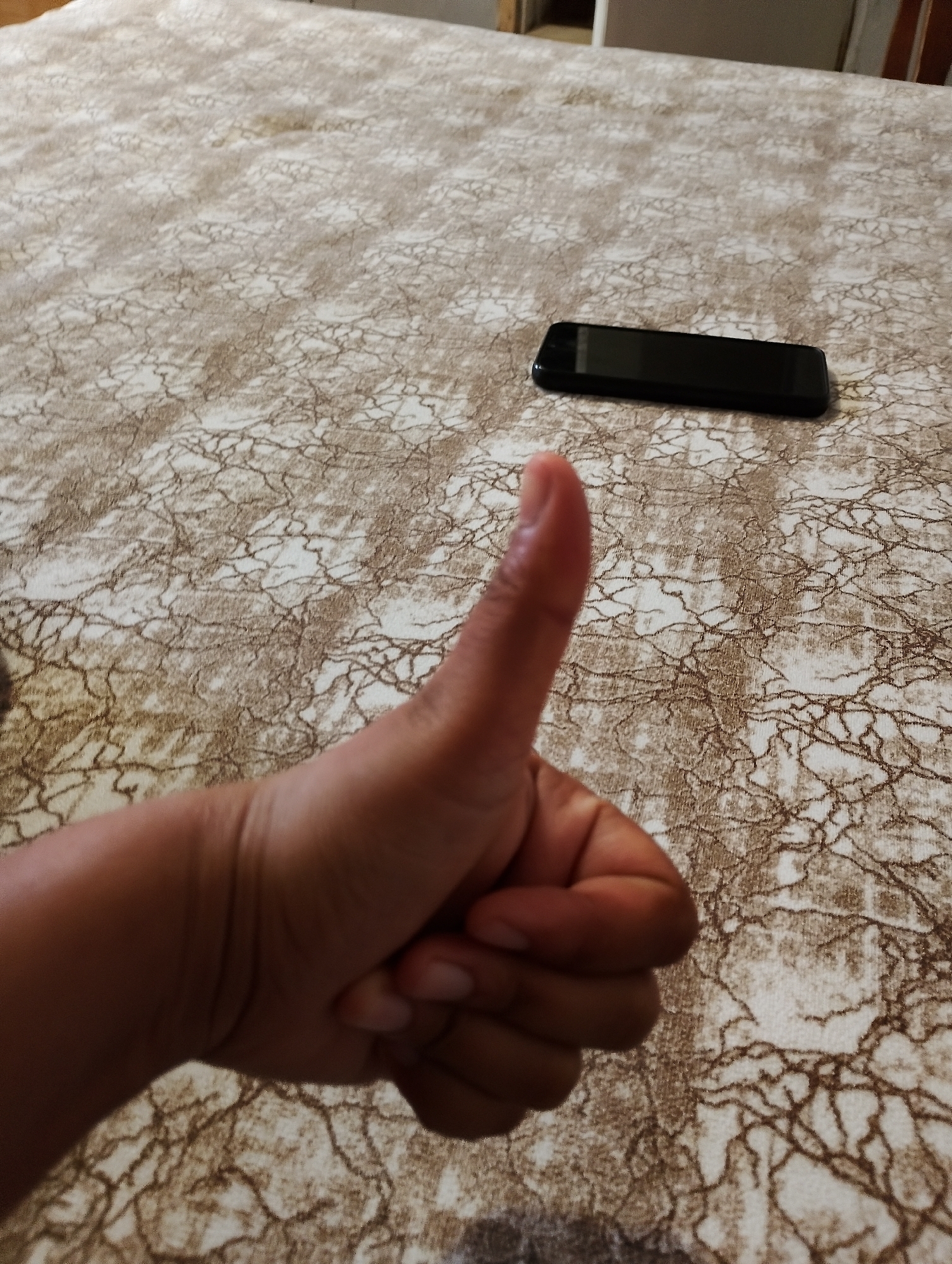 A hand is shown giving a thumbs-up gesture in front of a patterned tablecloth. A black smartphone is lying on the tablecloth to the right of the hand. The background is blurred, showing a table and possibly chairs. The scene appears to be indoors. The location is Yambol, Bulgaria.