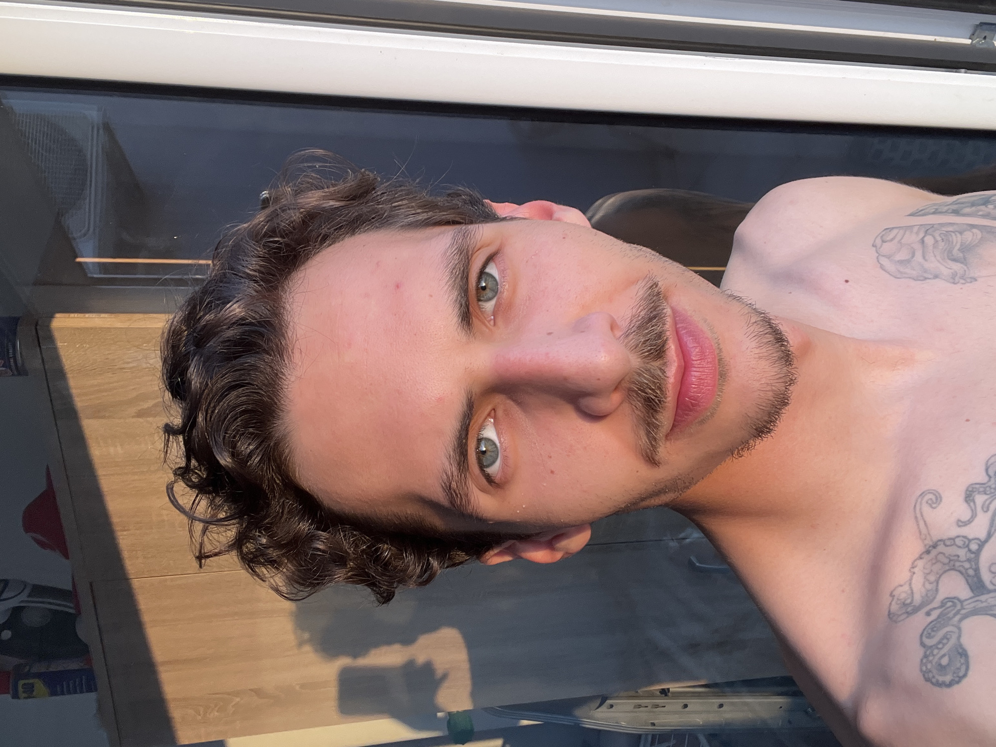 A close-up, angled shot shows a shirtless man with curly brown hair, hazel eyes, and a light mustache. He is looking directly at the camera. Parts of tattoos are visible on his right shoulder and chest. He is indoors, standing near a window with a reflective surface. The reflection reveals what appears to be part of a room with a wooden floor and some objects on a shelf or table. Without additional context, it's impossible to pinpoint a specific location in Rafailovići, Montenegro.