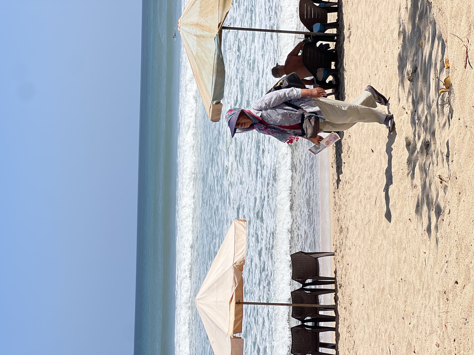 The image depicts a sunny day at Kuta Beach, Indonesia. A woman wearing a hat, a patterned shirt, and light-colored pants walks along the sandy shore. She carries a small bag across her body and holds some papers in her hand. In the background, the ocean features rolling waves and a clear blue sky. Two beach umbrellas provide shade, with several empty plastic chairs placed beneath them. Further in the background, two individuals are seated under an umbrella, one appearing to be shirtless. A few surfers can be seen in the water. The sand is light brown with scattered dry leaves and debris. The overall scene conveys a relaxed beach atmosphere.