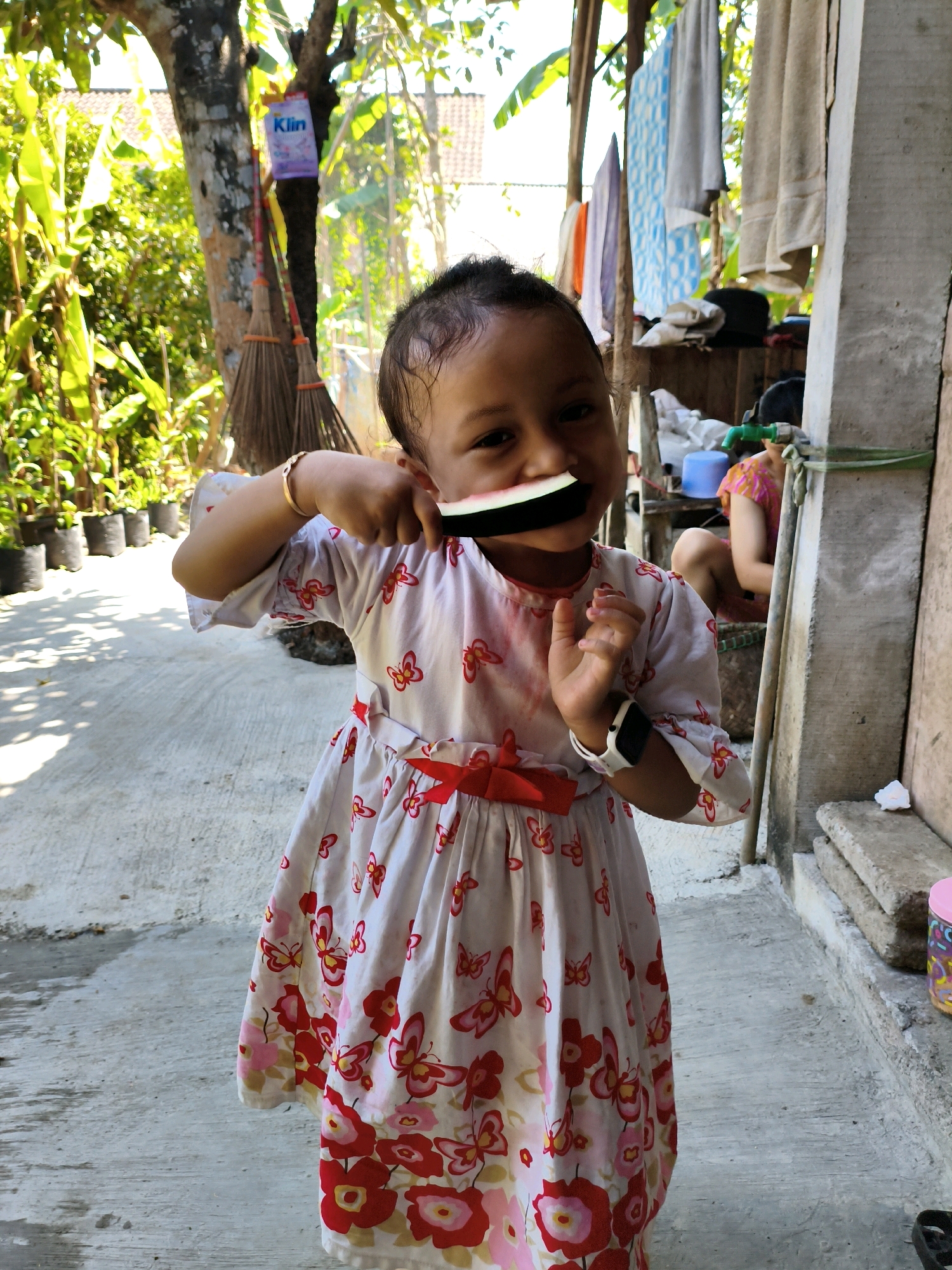 A young child, appearing to be of Indonesian descent, stands in an outdoor setting with a concrete ground and a building in the background. The child is wearing a white dress with a red butterfly and floral pattern, and a red sash. They are holding a slice of watermelon up to their mouth, with their hands positioned around it. The child has a bracelet on their left wrist and a watch on their right wrist. In the background, laundry is hanging on a line, and there are plants in pots. To the right, another person is visible, partially obscured, wearing a pink and orange patterned shirt. The scene suggests a residential area, possibly a yard or alleyway, in the village of Karangtalun, Indonesia.