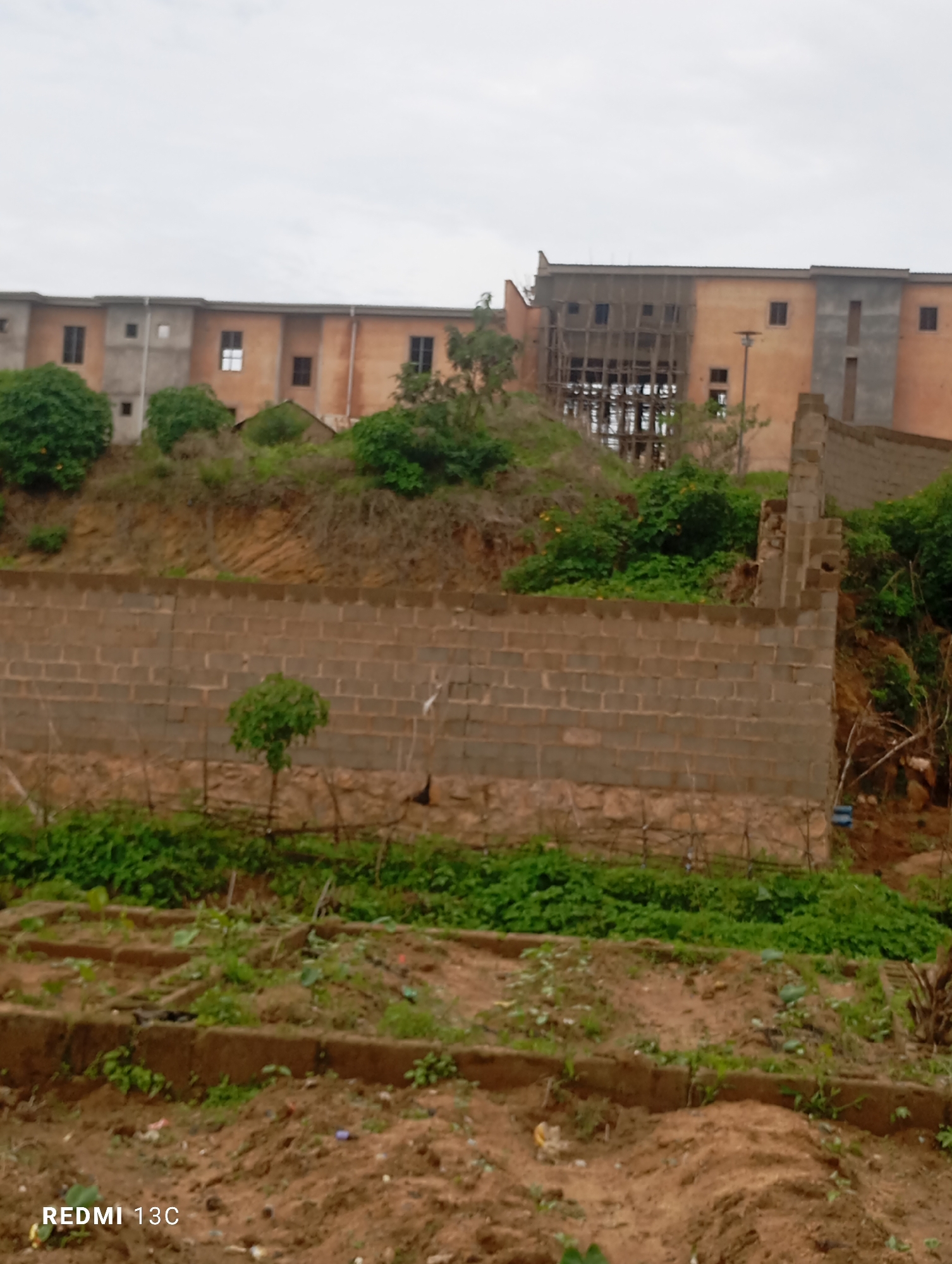 The image depicts a construction site in Jos, Nigeria, featuring a multi-story building under construction. The building exhibits varying stages of completion, with some sections appearing to have a finished facade in shades of orange and grey, while others are unfinished, revealing exposed concrete and wooden scaffolding. In the foreground, a partially constructed brick wall runs horizontally across the frame, with vegetation growing around and behind it. Beyond the wall, a dirt area with raised garden beds and sparse greenery suggests an agricultural or garden space. The background is dominated by the imposing structure and a cloudy sky. The overall scene conveys a sense of ongoing development and unfinished infrastructure within an urban environment.