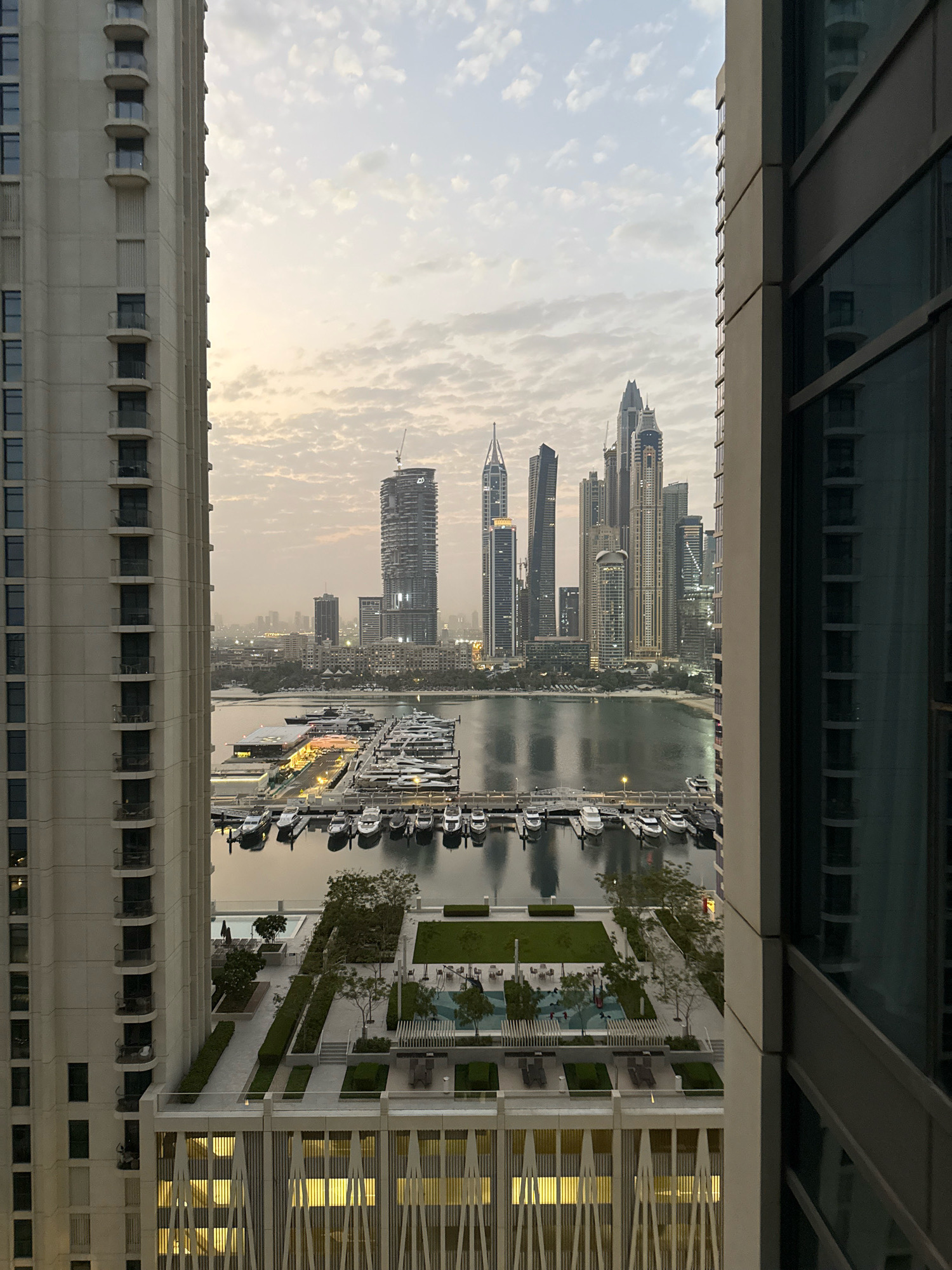 The image captures a panoramic view of the Dubai Marina, United Arab Emirates, at dusk. The scene is dominated by towering skyscrapers, characteristic of the city's modern architecture, juxtaposed with a serene waterfront. In the foreground, two large residential buildings frame the view, with balconies and windows visible on the left structure. The right building's dark, reflective glass facade creates a stark contrast.
The middle ground features the Dubai Marina, a bustling hub with numerous yachts and boats docked. The water reflects the soft evening light and the surrounding cityscape. Beyond the marina, a sandy beach area is visible, leading to more urban development.
The background is a cityscape of iconic Dubai skyscrapers, including those in the Marina and Jumeirah Beach Residence areas, illuminated against a hazy, pale sky. The overall atmosphere is one of urban sophistication and tranquility, with the city lights beginning to twinkle as night approaches. No people are clearly visible in the image.