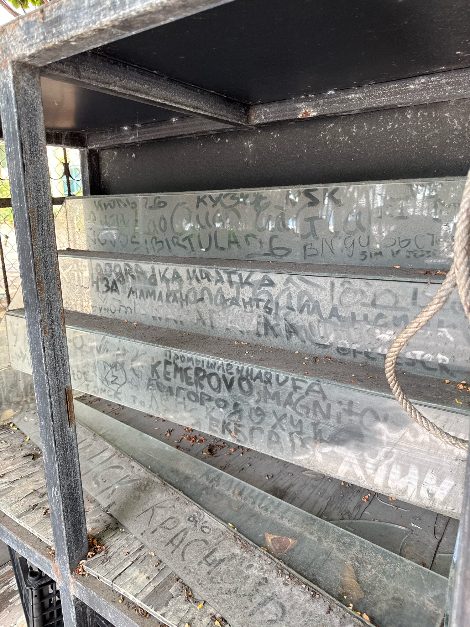 The image shows a set of glass shelves covered in graffiti. The shelves are part of a dark metal structure, possibly a display rack or shelving unit. Dust and fallen leaves are visible on the shelves. A thick rope is hanging down the right side of the image. Some of the graffiti appears to be in Cyrillic script, with phrases like "KEMEROVO" and what looks like "KRASIVYY" (beautiful). The overall impression is of an abandoned or neglected area. Based on the Cyrillic graffiti, it is unlikely that this location is in Nha Trang, Vietnam, as this script is not used in Vietnam.
