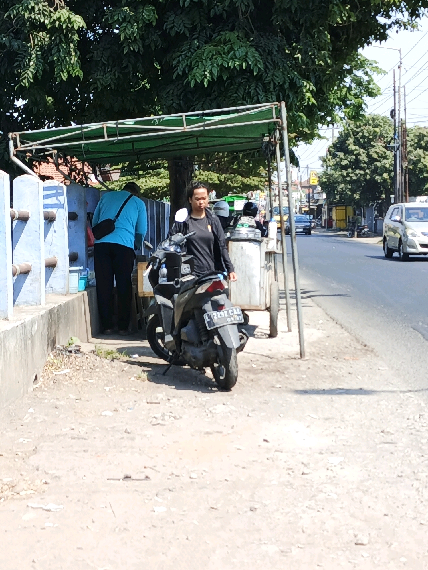 A medium shot captures a street scene in Ketegan, Indonesia, on a bright, sunny day. Two individuals are present. In the foreground to the left, a person wearing a bright blue long-sleeved shirt and black pants stands with their back to the camera, a black bag slung across their shoulder. To the right, a person in a black t-shirt and black pants stands facing left, looking towards the first individual. They are positioned next to a motorcycle. The motorcycle is dark-colored, parked with its front wheel turned slightly to the left. Behind the individuals, a simple stall with a green canopy supported by metal poles is set up. A cart-like structure, possibly part of the stall, is visible. The ground is a mix of gravel and dirt. A blue, tiled wall is on the far left. The background shows a paved road with passing vehicles, including a white car on the right and a dark car further down the road. Trees with dense green foliage are prominent in the upper portion of the image, and utility poles and wires are visible against the sky. The scene appears to be an outdoor food stall or a small roadside vendor.