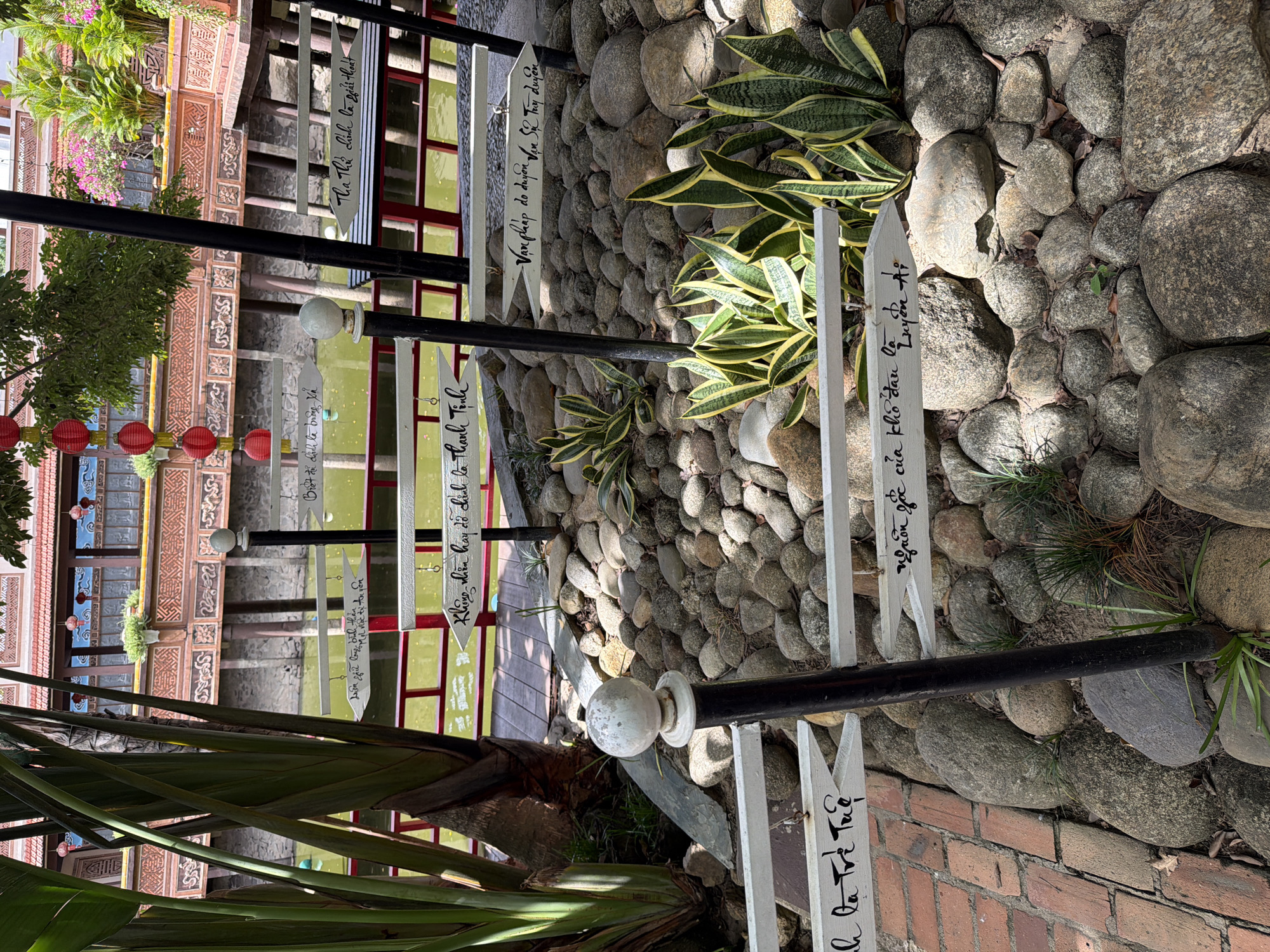 The image depicts a garden scene in Hoi An, Vietnam, featuring a series of white directional signs with handwritten Vietnamese text. The signs are mounted on a black pole and point in various directions, indicating different destinations or concepts. In the background, a traditional Vietnamese building with red tile roofing and ornate wooden detailing is visible, overlooking a body of water. Red lanterns are suspended from the building's eaves. The foreground is filled with various sizes of river stones and a few small green plants, including a Snake Plant with variegated leaves. To the left, a large fan-shaped palm tree stands prominently. The ground in the lower left corner is paved with red bricks. The overall impression is a tranquil outdoor space with cultural and natural elements.