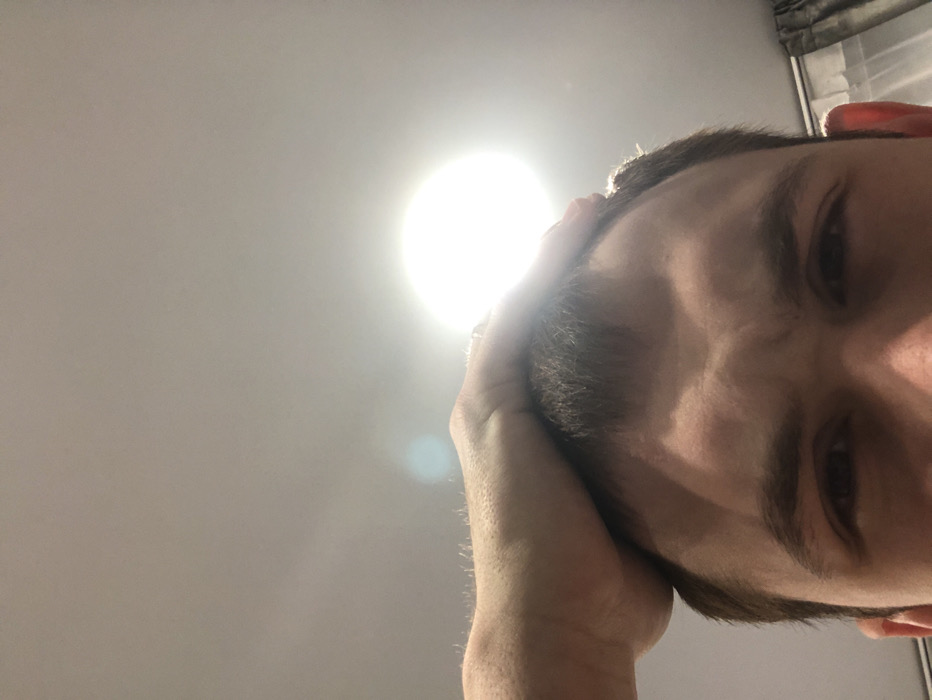A man's face dominates the right of the frame, with his dark hair and furrowed brows in sharp focus. He appears to be resting his head in his hand. To the left, a bright, circular light source washes out the background with intense luminosity, creating a strong contrast. The angle is a low-angle perspective, looking up at the man's face and the light.