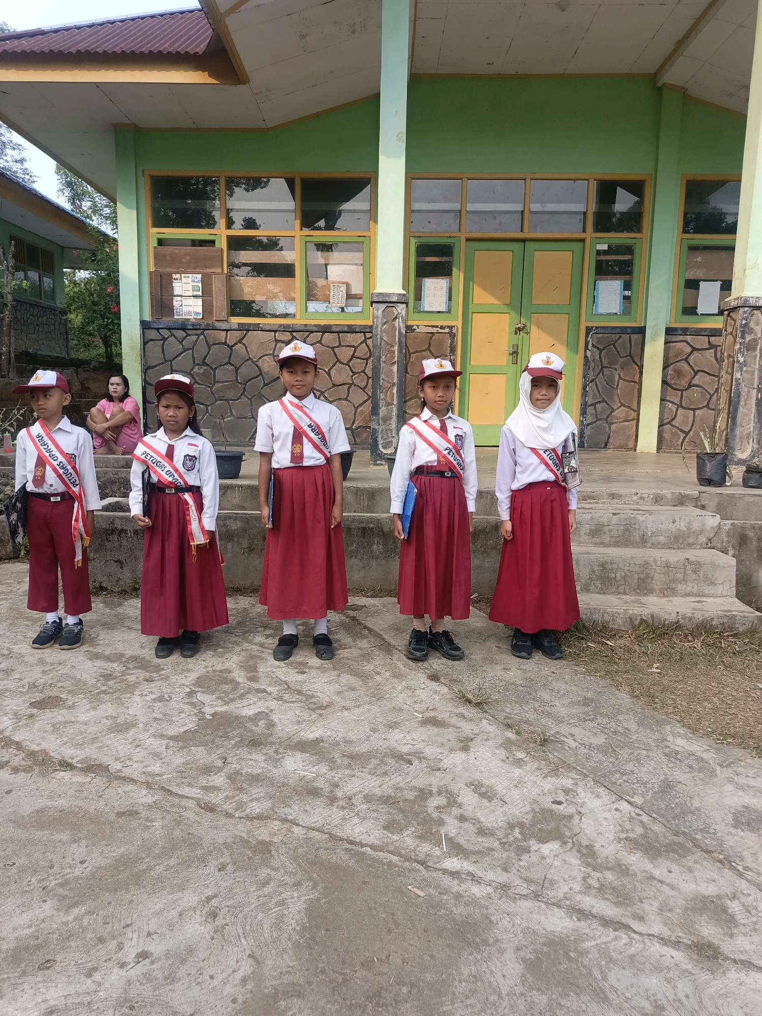 Five children, three boys and two girls, stand in a line on a concrete walkway in front of a building. They are all wearing school uniforms consisting of white shirts, red skirts or pants, and red sashes. Each child also wears a white cap with a red emblem. The building behind them is painted green and yellow with a stone facade. In the background, a woman sits on some steps. The scene appears to be a school in Taba Tinggi, Indonesia.