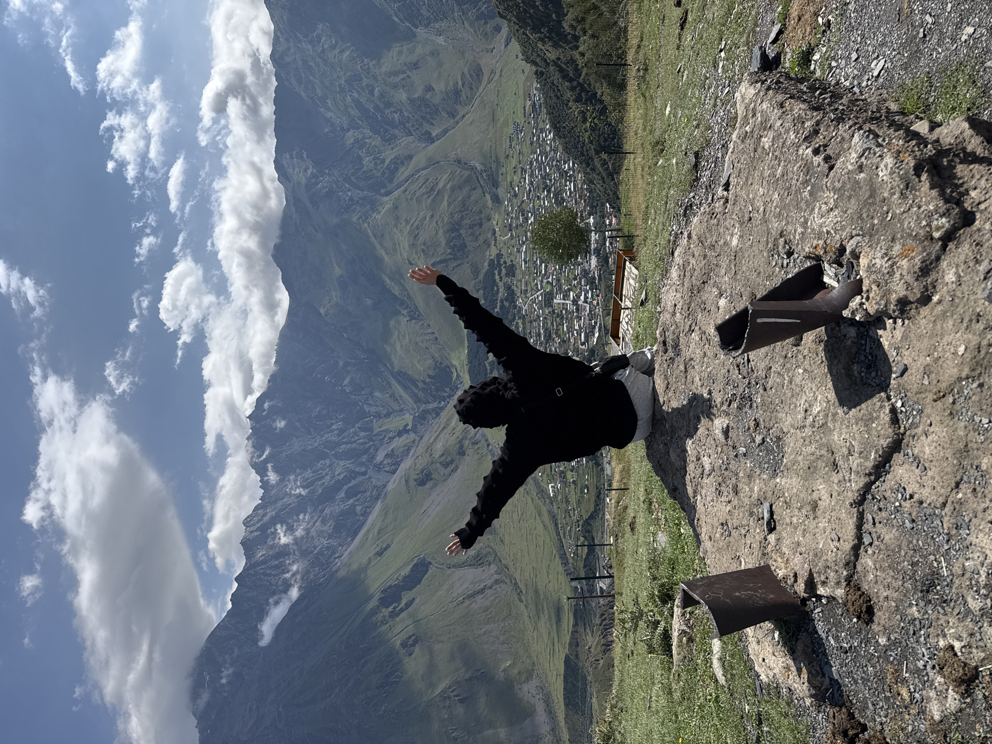 A person wearing a black hooded jacket and light-colored pants is sitting on a concrete structure, facing away from the camera with arms outstretched. The background features a vast, mountainous landscape with a town nestled in the valley below. The sky is partly cloudy, with dramatic formations of white and gray clouds above the rugged, green mountains. The foreground shows the textured surface of the concrete structure with metal pieces embedded in it, surrounded by dry grass and gravel. The town appears to be Stepantsminda, Georgia, known for its stunning mountain views.