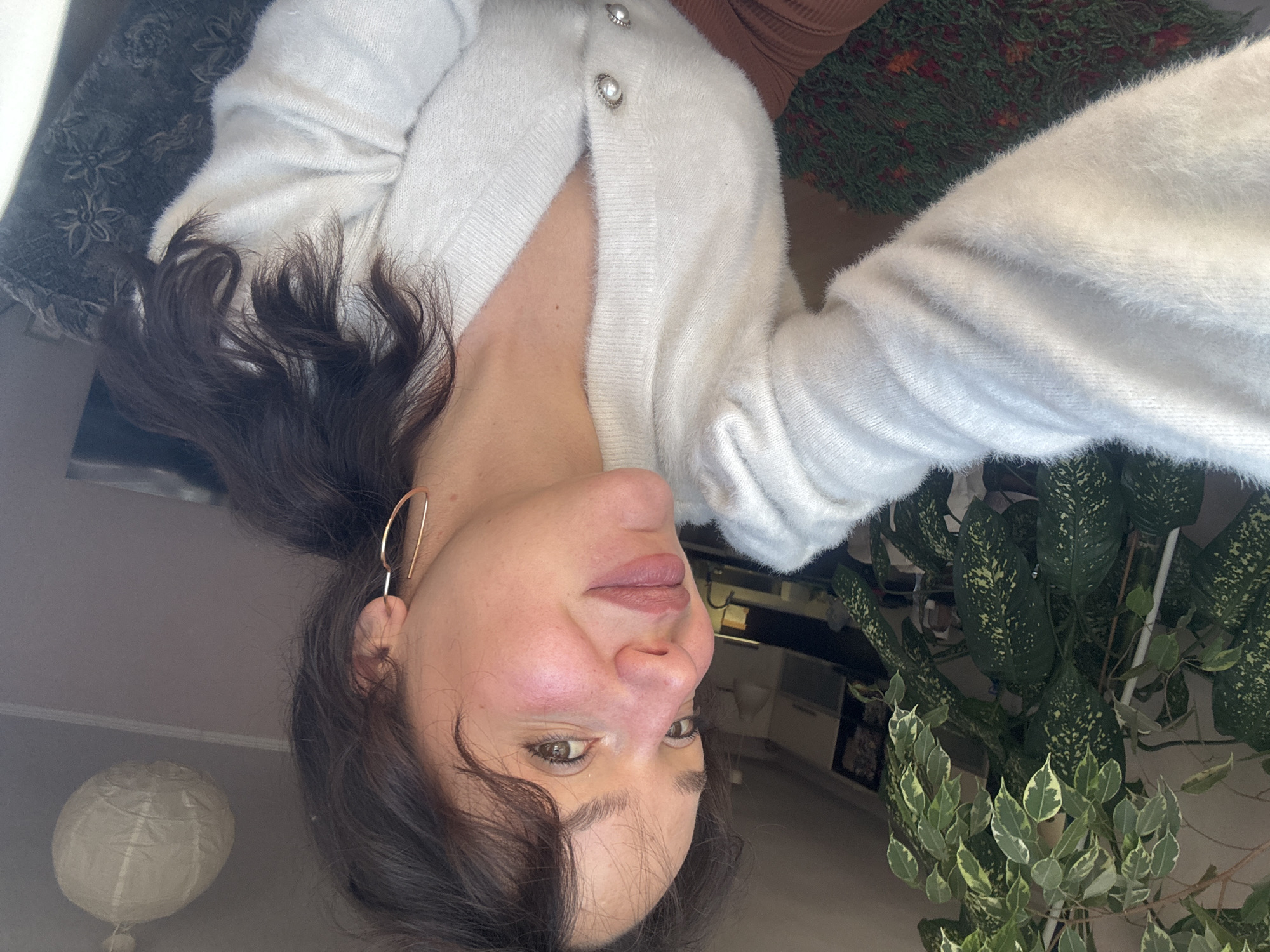 A selfie portrait shows a woman indoors, likely in a personal residence. She is oriented upside-down in the frame. She is wearing a white, fuzzy cardigan with pearlescent button details. She has dark brown hair with gold hoop earrings. A houseplant with green and white variegated leaves is visible in the bottom right corner. Above the woman, there is a glimpse of a floral patterned pillow. A white paper lantern is situated in the bottom left. The background suggests a modern interior with light walls and glimpses of furniture. The image does not provide information related to Burgas, Bulgaria.