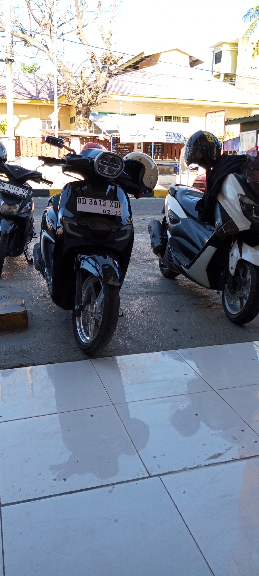 The image depicts a street scene in Assorajang, Indonesia, featuring three parked scooters. The scooter in the foreground, a black model, displays a license plate reading "DD 3612 XDF" with "02.31" below it. Two helmets are visible, one beige and one black, placed on the handlebars of the scooters. To the left of the central scooter, another black scooter is partially visible with a license plate reading "DDW 3315 NS" and "05-20." A white and gray scooter is parked to the right. The background reveals buildings with yellow and white facades, a large tree with bare branches, and power lines. The ground is paved with tiles in the foreground and textured concrete behind the scooters.
