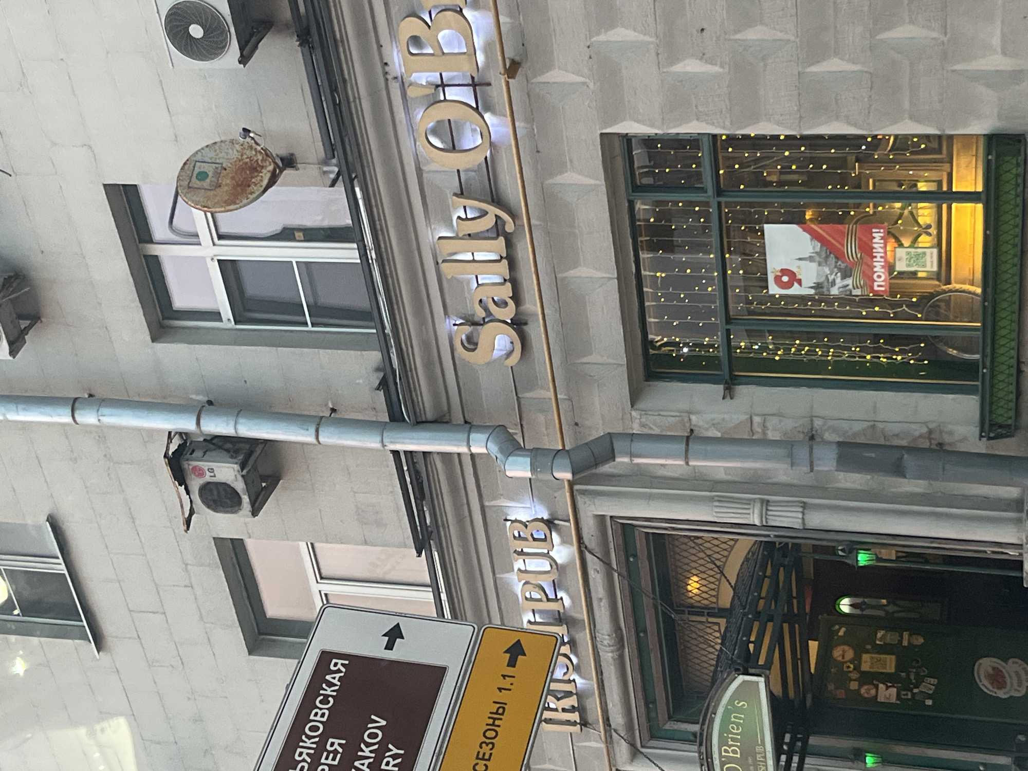Here's a forensic description of the image:
**Location:** Moscow, Russia
**Scene:** An exterior shot of two establishments in Moscow.
**Establishments:**
* "Sally O'Brien's Irish Pub" - signage visible above its entrance.
* A shop/business next door labeled "Sally O'B." The sign is above a window decorated with lights and a poster that contains the word "ПОМНИМ!" (meaning "Remember!" in Russian).
**Objects:**
* Street signs: A street sign with the word "ЛЯКОВСКАЯ" and another reading "СЕЗОНЫ 1.1."
* Air conditioning units.
* Pipes running along the building's facade.
**Environment:**
* Stone or concrete exterior building.
* Windows, visible lighting within the buildings.