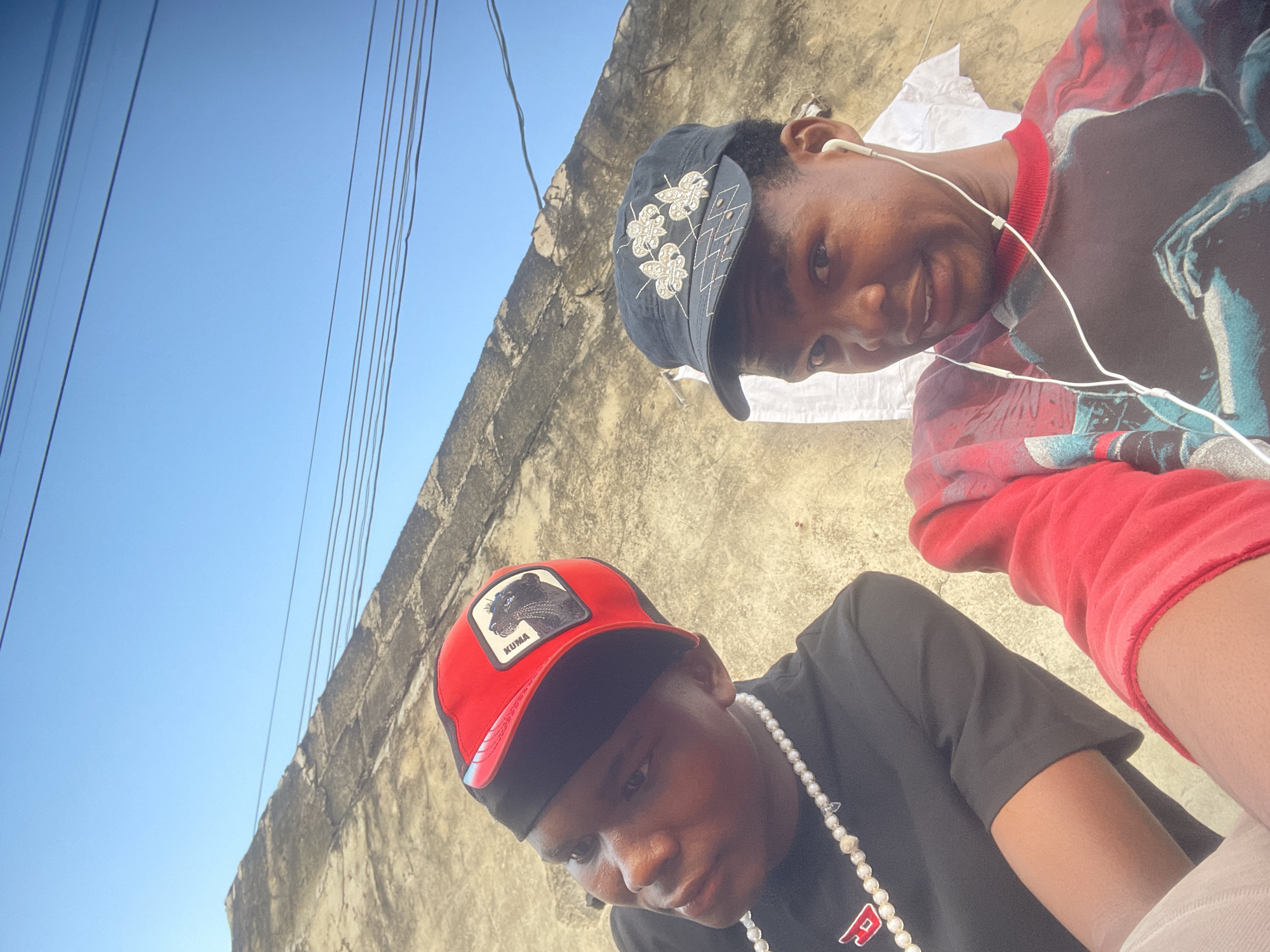 In Ado, Nigeria, two young men are pictured outdoors, seated and looking towards the camera. The man on the left wears a black t-shirt with a red logo and a red cap with a black brim featuring a graphic of a black panther. He also wears a pearl necklace. The man on the right wears a black cap adorned with decorative flowers and a long-sleeved shirt with a graphic print. He has white earbuds in. They are positioned in front of a rough, textured wall, possibly a building exterior, under a clear blue sky. Power lines are visible strung across the top of the image.