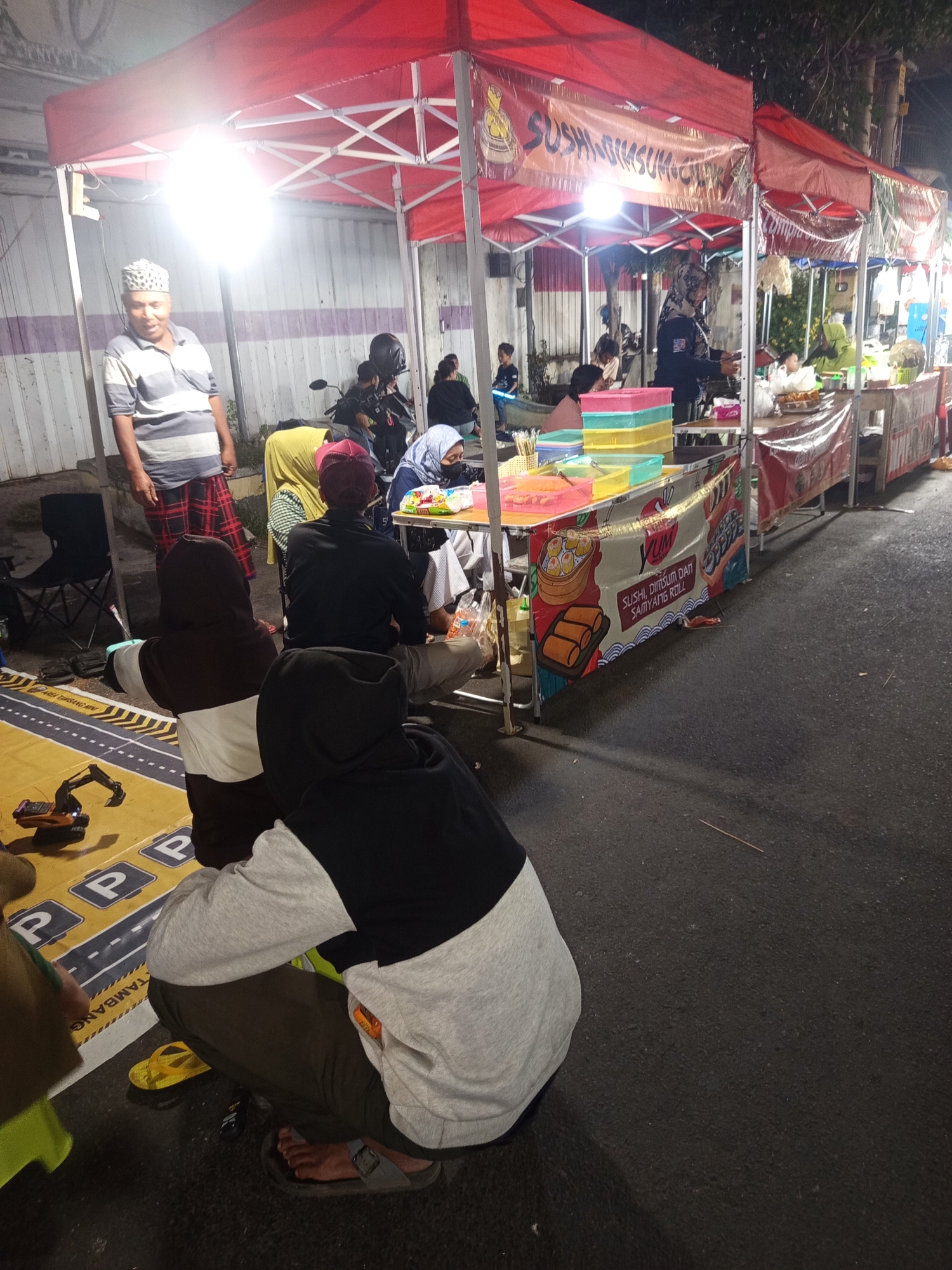 The image captures a nighttime scene at a street food stall in Ardirejo, Indonesia. A red tent provides cover for a food vendor with various dishes displayed on tables. Several people are gathered around, some ordering or eating, while others are seated. A man in a striped shirt and a cap stands near the entrance of the tent, appearing to be a vendor or attendant. In the foreground, two individuals are seated on the ground, one wearing a gray and black hooded sweatshirt. The ground is paved with asphalt and decorated with yellow and white street markings and parking symbols, suggesting the stall is set up on a roadside or designated area. Yellow flip-flops are visible near the seated individuals. The overall atmosphere suggests a busy evening at a local market or food court.
