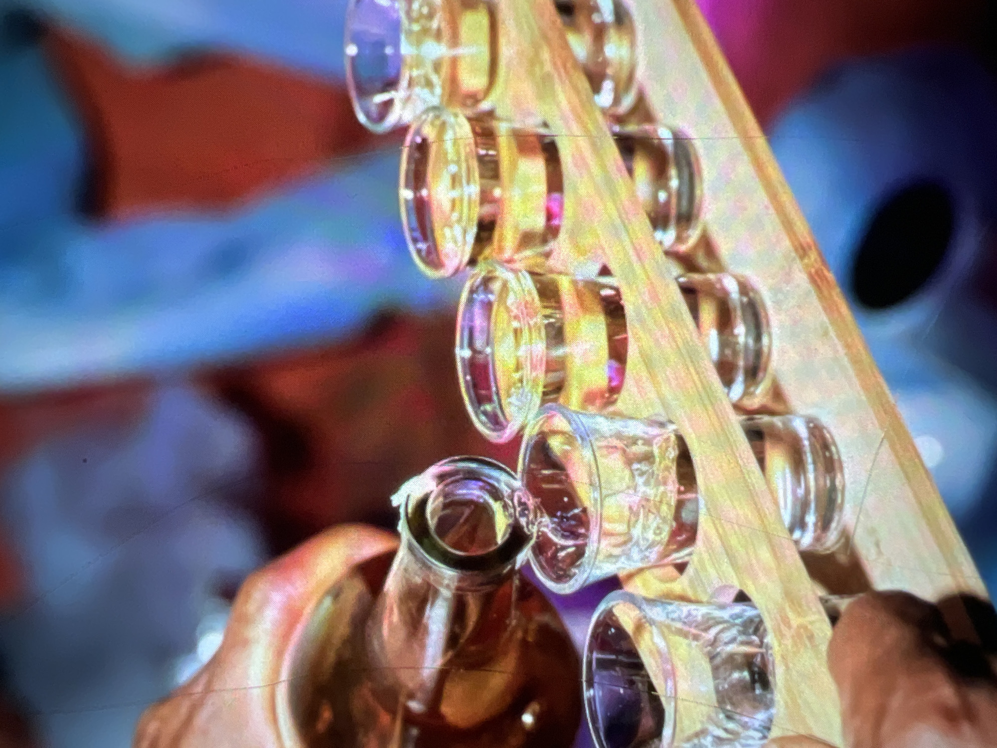 In the city of Lagos, Nigeria, a hand is pouring a golden liquid from a glass bottle into a series of small shot glasses lined up on a wooden tray. The glasses are partially filled, with the liquid catching the light and showing distinct amber hues. In the background, the scene is blurred, with suggestions of colorful, abstract shapes that could indicate a lively environment, possibly a bar or a party. The focus is entirely on the act of pouring and the presentation of the drinks.