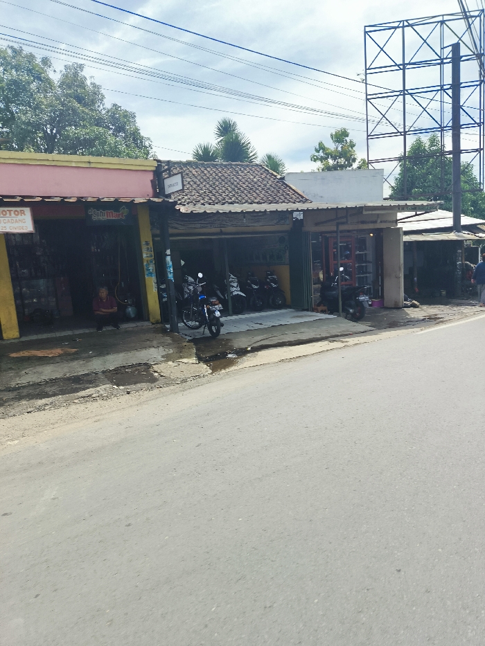 The image shows a street scene in Ciwidey, Indonesia, located at the coordinates -7.105 latitude and 107.458 longitude. The scene is set in front of several shops. On the left side of the street, a yellow building has a sign that reads "MOTOR CADANG ONDES". Further down the street, a small shop is seen with its garage doors open and various motorcycles parked inside. A man in a dark shirt and pants stands in front of the shop. Next to the shop is a tall, metal tower with a criss-cross pattern. On the right side of the street, a small building is visible, with a sign reading "Sari Mart". The street is paved with gray asphalt, and the background features green foliage and a clear, partly cloudy sky.