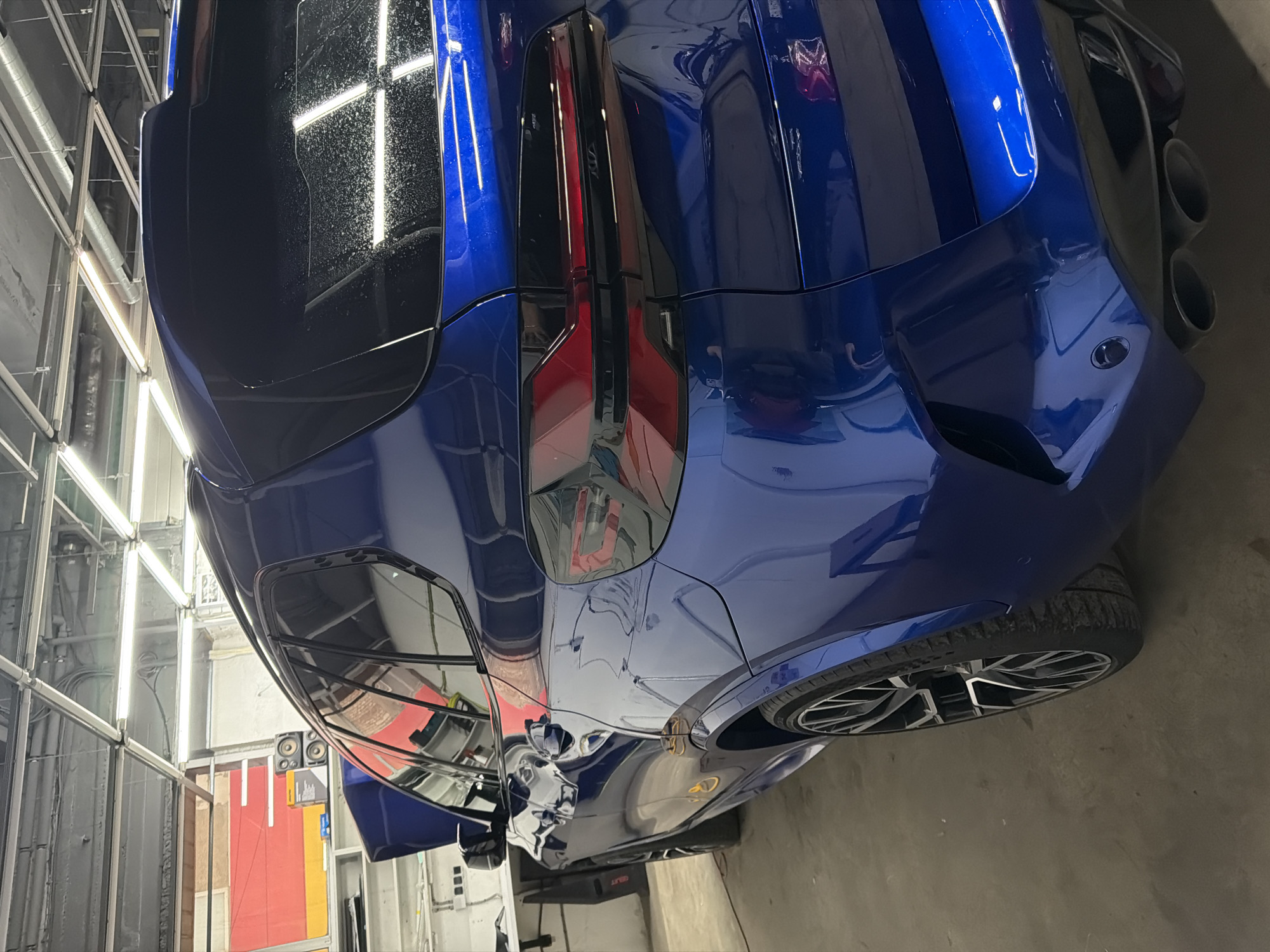 The image depicts the rear of a bright blue car, likely a sports car, from an elevated perspective. The car features dark-tinted windows, distinctive rear lights with red accents, and prominent exhaust pipes. The car is parked indoors on a concrete floor within what appears to be a detailing or maintenance facility, given the presence of overhead fluorescent lights and adjacent equipment.
The location is difficult to determine based only on the picture and without additional information. It could be Krasnoyarsk, Russia, but that cannot be independently verified.