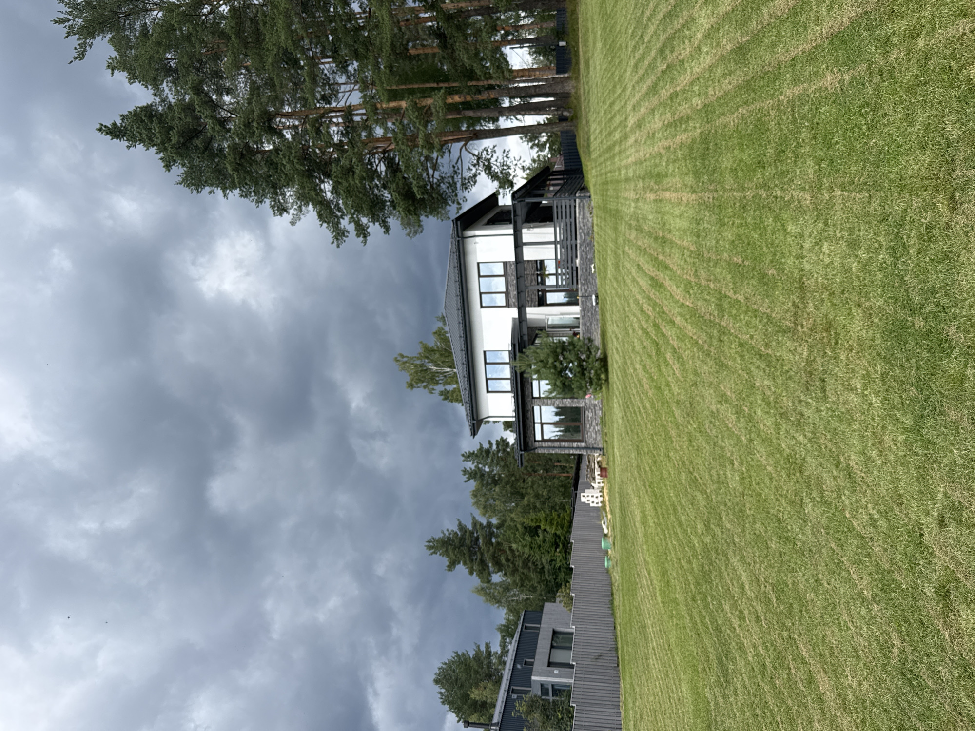 The image depicts a modern two-story house with white and gray facades, a dark roof, and large windows. It is situated in a suburban or rural setting, surrounded by a neatly manicured green lawn with visible mowing patterns. Tall pine trees and other deciduous trees are present to the right and in the background. A wooden fence separates the property from a neighboring area, where another contemporary house with a gray exterior and darker roofing is visible. The sky is overcast with dark, heavy clouds, suggesting an impending storm or gloomy weather. There are no people or animals visible in the scene. The location is identified as Mars, Russia.