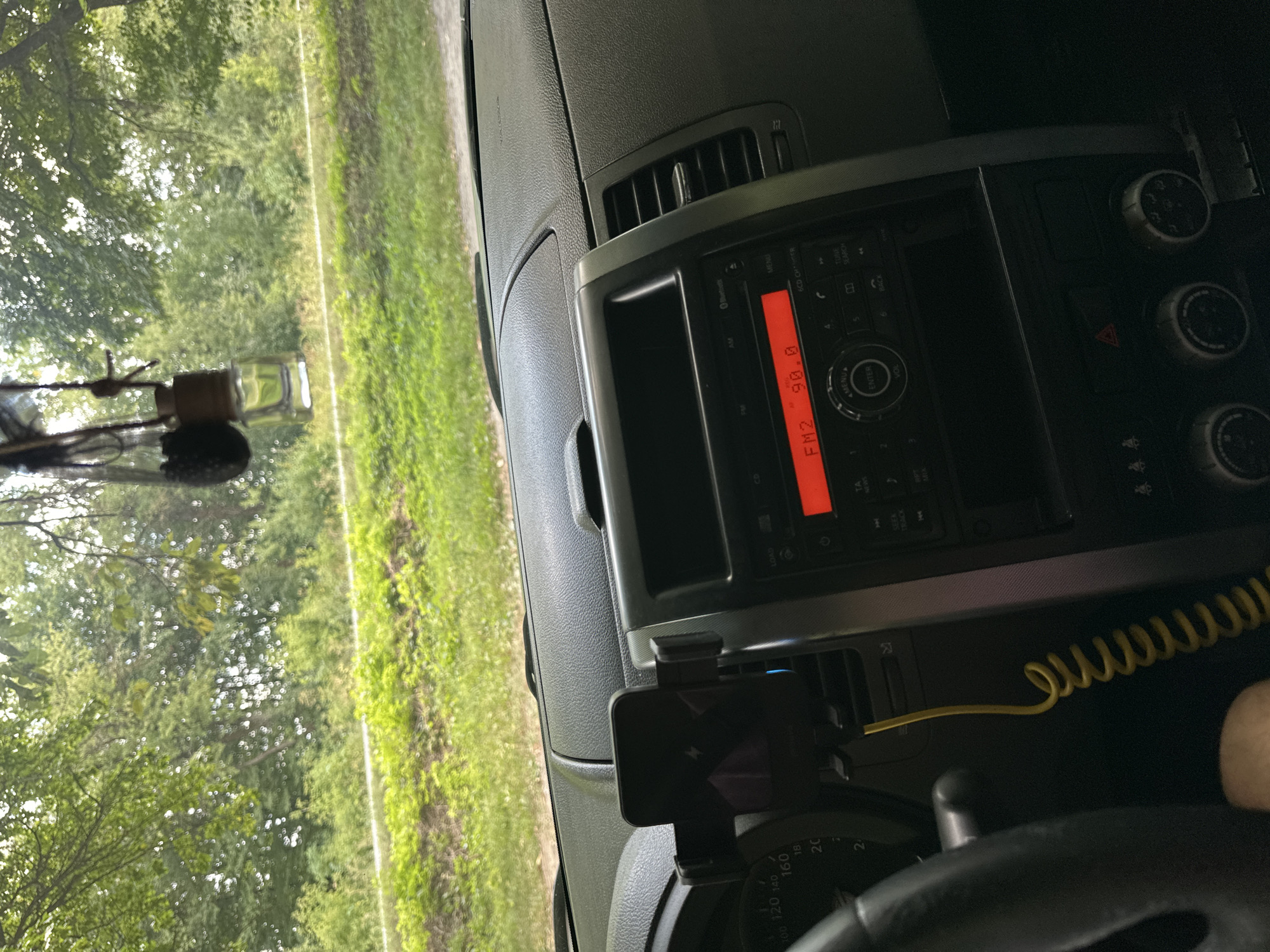 The image shows the interior of a car, with a view through the windshield to a rural, green landscape. The car's dashboard is visible, featuring a car stereo displaying "FM2 90.0", a phone mounted on a holder, and other controls. A yellow coiled cable extends from the dashboard. Outside the car, trees and vegetation are abundant, suggesting a natural or undeveloped area. No specific urban features of Shatava, Ukraine, are discernible from this view.