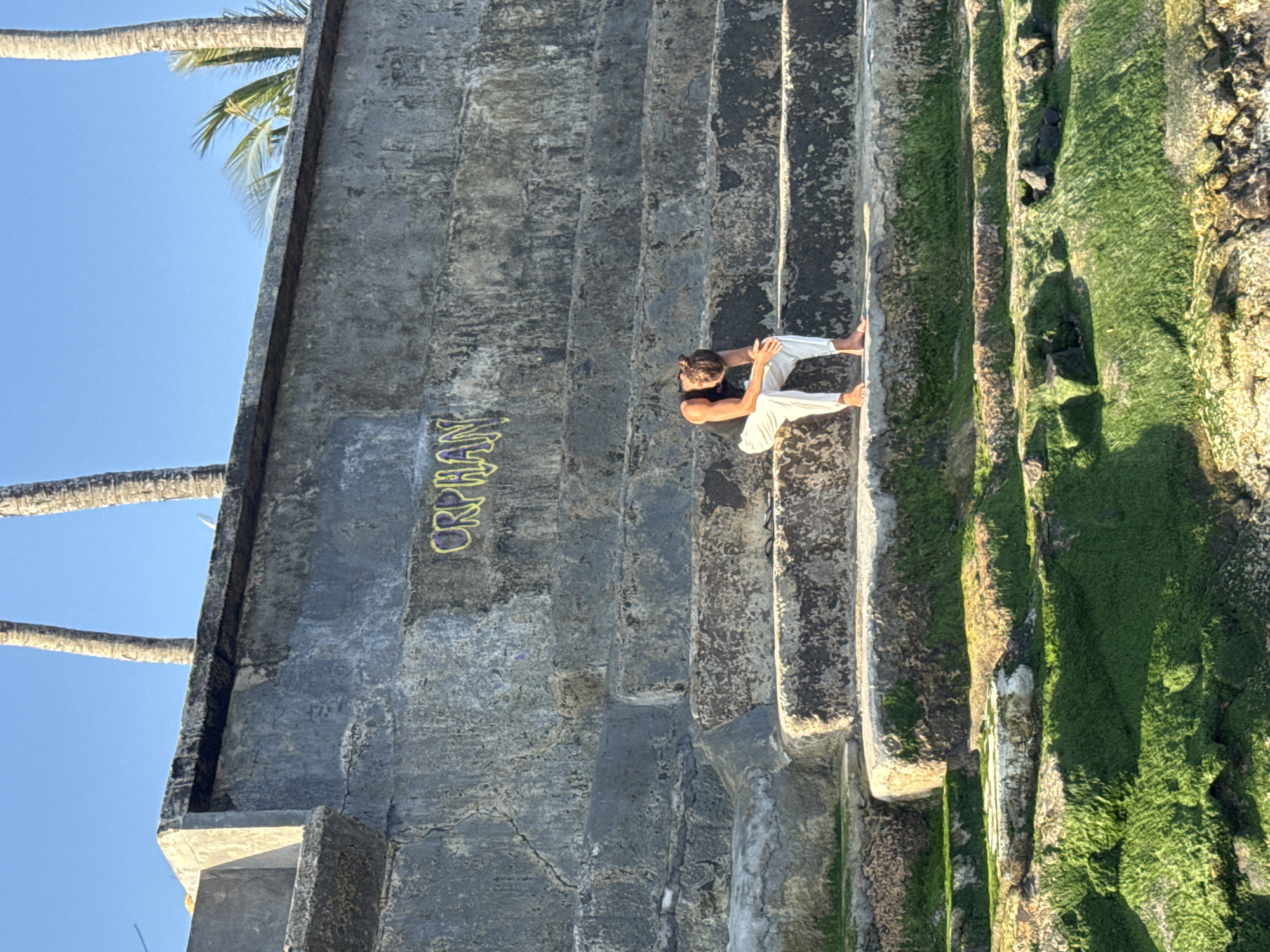 A young man in white pants and a dark tank top sits on a wide stone step, his head bowed and hands clasped. He is positioned on a set of weathered, concrete stairs that descend towards a body of water. The stairs are partially covered in vibrant green moss. Above the stairs, on a concrete wall, graffiti in yellow spray paint reads "ORPHAN." In the background, palm trees are visible against a clear blue sky. The scene appears to be outdoors in a coastal or waterfront area. The image was taken in the city of Mengwi, Indonesia.