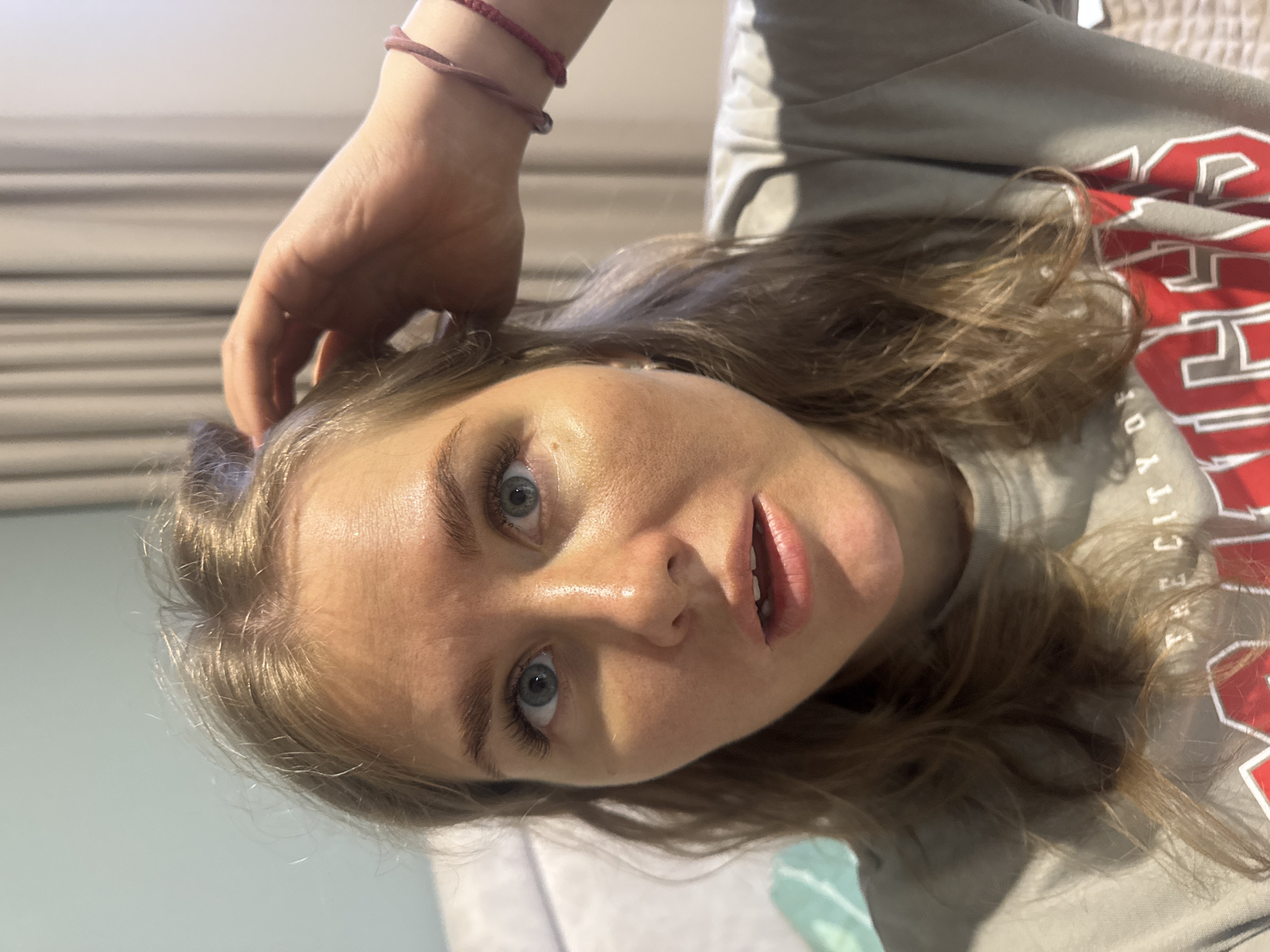 A young woman with light brown hair and blue eyes is shown close-up. She is wearing a gray sweatshirt with red lettering that reads "THE CITY OF..." and what appears to be "DY" followed by other letters that are illegible. Her left hand, adorned with two thin red bracelets, is touching her hair. The background is a blurred view of a wall with horizontal lines. The location is unknown, but the city Ферма, Russia is mentioned. There are no actions or interactions visible except the woman's hand touching her hair.