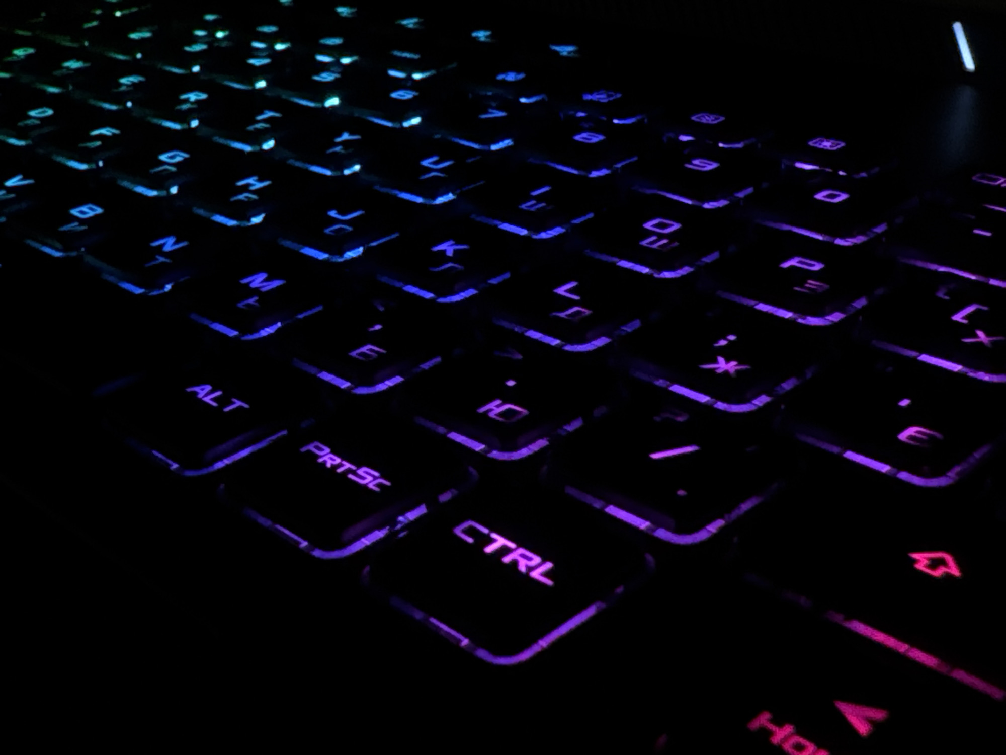 The image is a close-up, low-angle, eye-level shot of a black keyboard with customizable RGB backlighting. The lighting is predominantly blue and purple, with some green visible in the upper left portion of the image. The keys are illuminated, making the lettering and symbols on them stand out against the dark keys. Several keys are visible, including what appear to be letters like 'N', 'M', 'K', 'L', 'P', 'O', 'I', 'U', 'Y', and function keys such as 'ALT', 'PRTSc', and 'CTRL'. The lighting is bright and vibrant, creating a futuristic or gaming aesthetic. The background is completely dark, with no discernible environment or location details. There are no people or any other objects visible in the frame besides the keyboard itself. The image is in focus, and the detail on the keys is sharp.