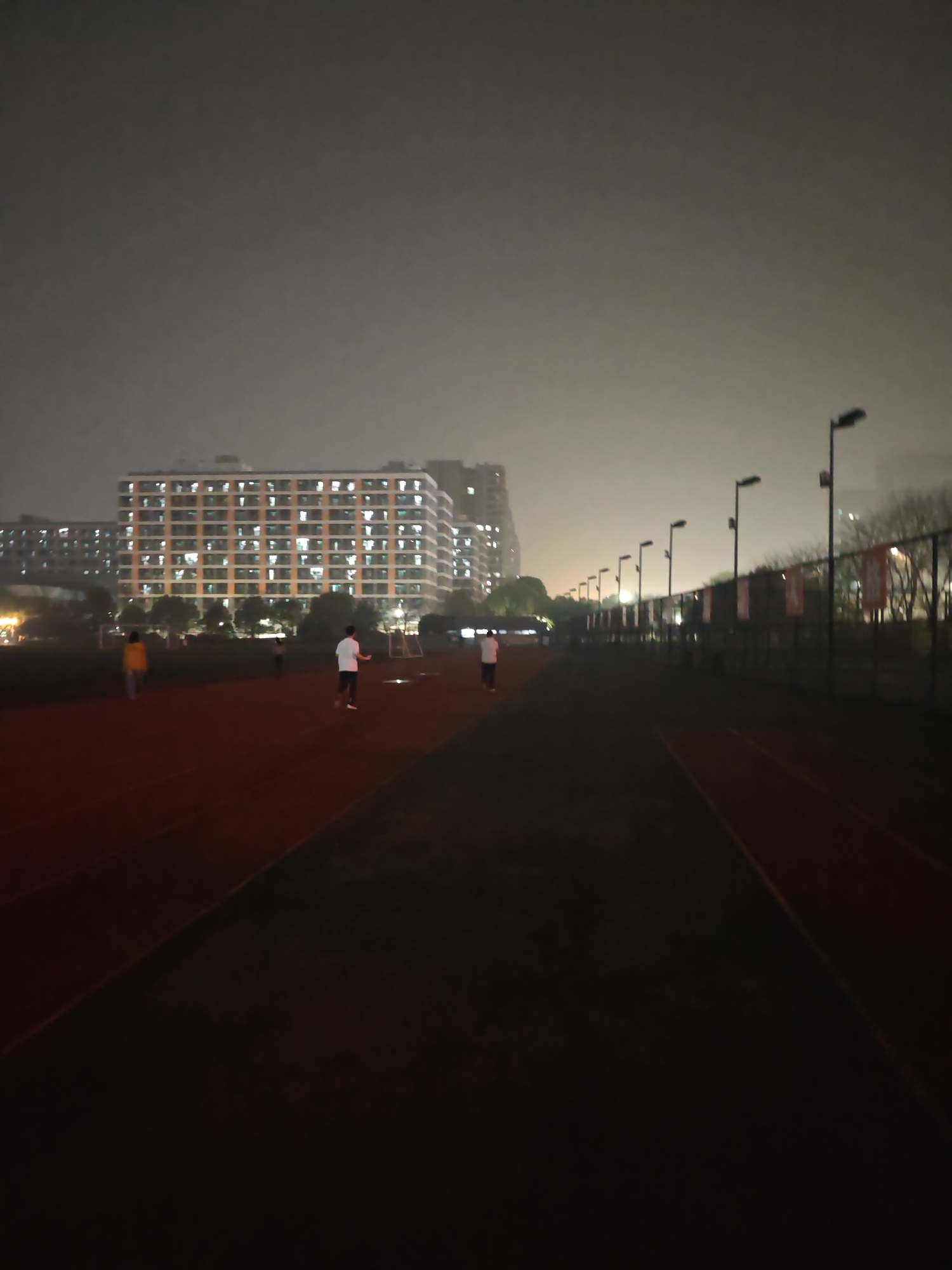 The image depicts a running track at night in Jiangning District, China. Buildings with illuminated windows are visible in the background. Several individuals are present on the track, including people walking and running. The environment appears foggy or hazy, with streetlights illuminating the path. A fence with flags runs along the side of the track.