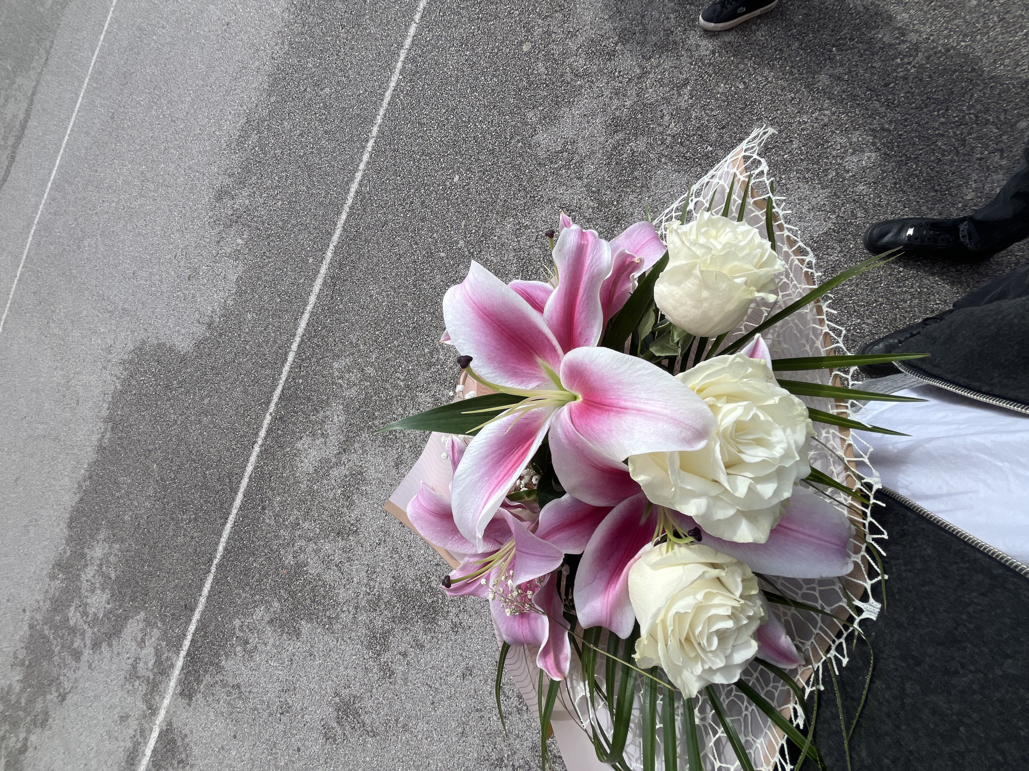 The image shows a bouquet of flowers, predominantly white roses and pink lilies, held above an asphalt surface. The ground appears to be marked with white painted lines, possibly indicating a road or parking area. Part of a person's clothing and footwear is visible in the top right corner, suggesting the bouquet is being held by someone standing on the asphalt. The environment seems to be outdoors, given the natural lighting. No specific landmarks are discernible, making precise location within Blagoevgrad, Bulgaria, impossible to determine from the image alone.