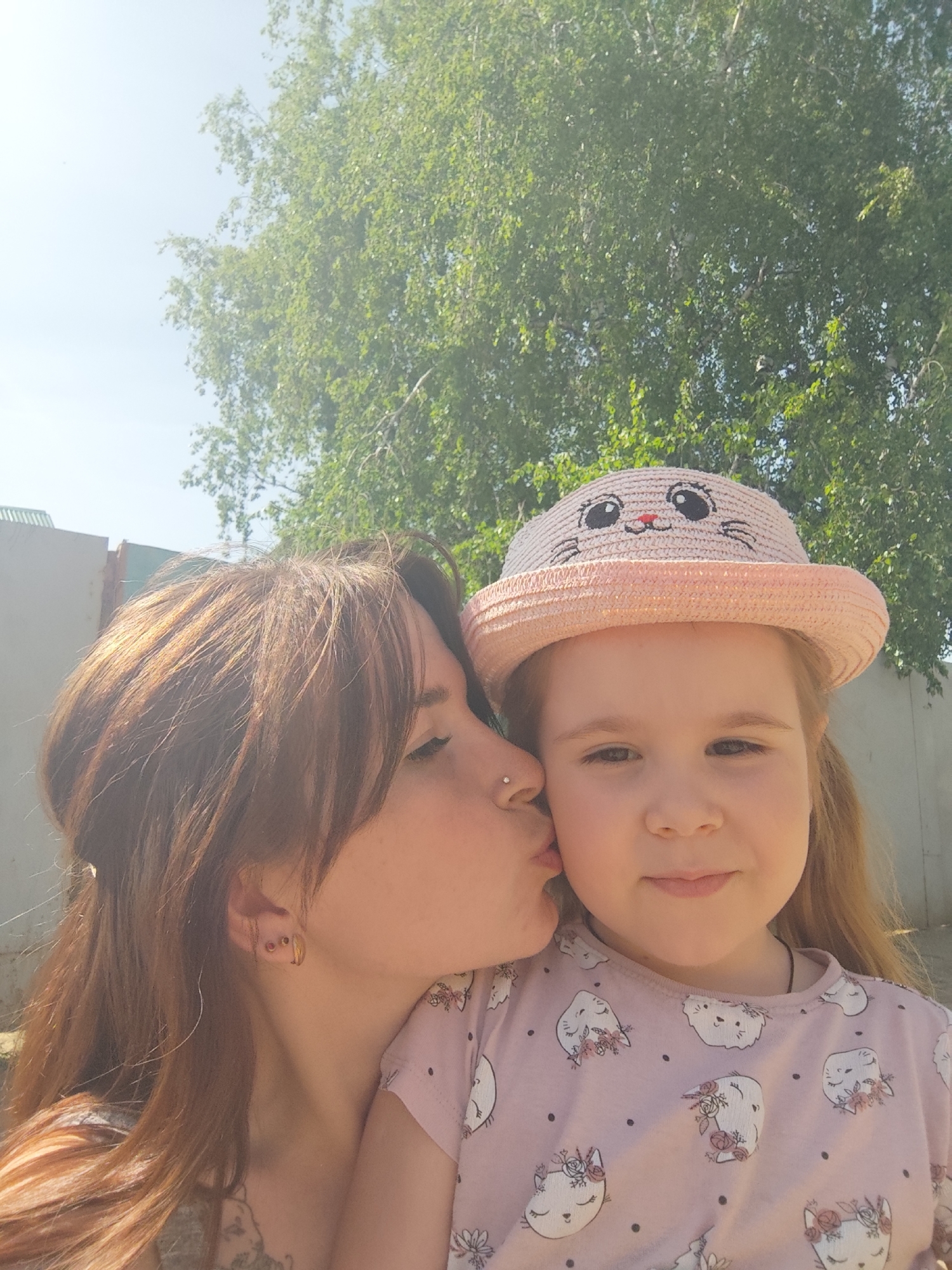 A woman is kissing a young girl on the cheek. The girl is wearing a pink shirt with a kitten pattern and a pink straw hat with a kitten face on it. The woman has brown hair and a nose piercing. They are standing outside in front of a tree. The location is Фоки, Russia. There is a fence in the background. The weather is sunny.