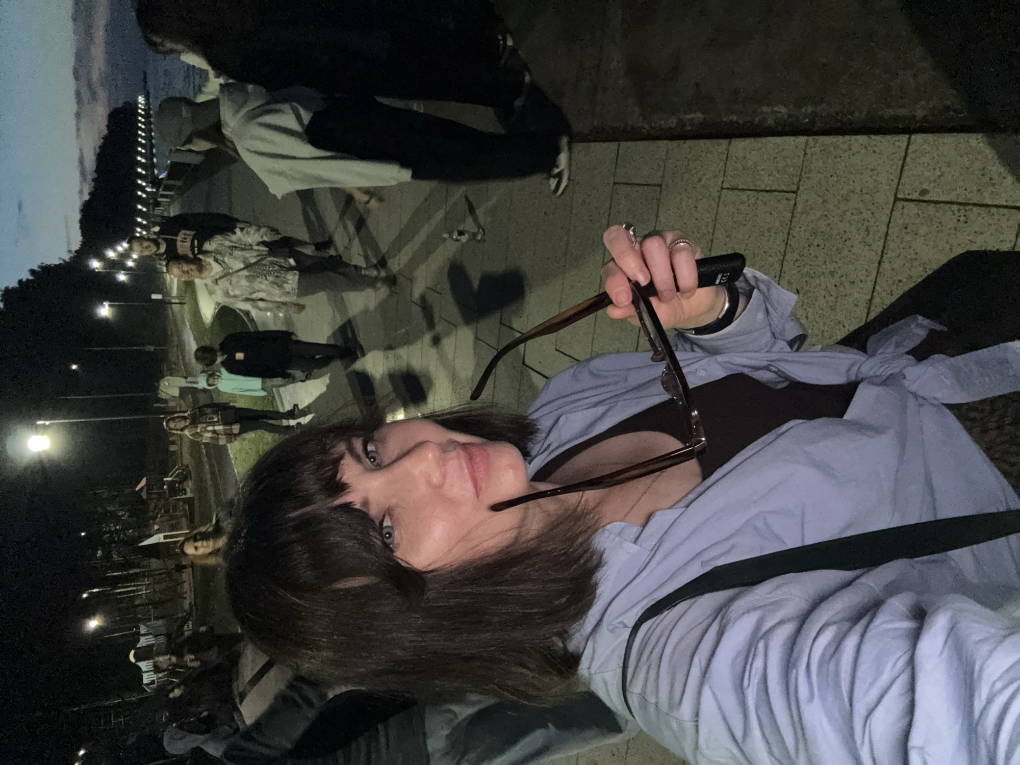 A woman is taking a selfie outdoors at night. She is in the foreground, holding a pair of glasses. She has short, dark hair and is wearing a light blue shirt. Behind her, a group of people are walking along a paved walkway next to a body of water. Streetlights illuminate the path. In the background, there is a playground and trees. The scene appears to be a waterfront promenade in Chaikovsky, Russia, given the presence of the Kama River and the typical urban recreational infrastructure.