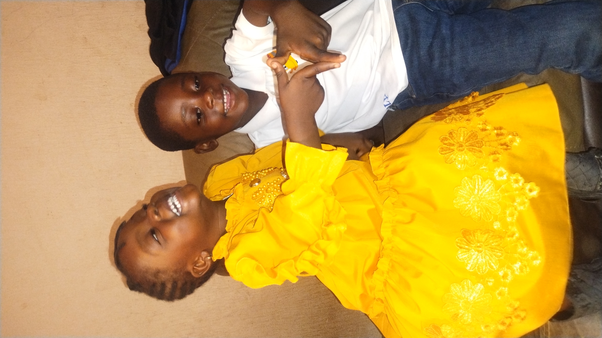 Two children are lying on a brown surface, possibly a couch or bed. The child on top is a boy wearing a white shirt and jeans. He is smiling and holding his hands up in a gesture. The child below is a girl wearing a bright yellow dress with floral embroidery and beaded embellishments. She is also smiling and looking upwards. The location is likely indoors. There is no readily identifiable information indicating this image was taken in Jos, Nigeria.